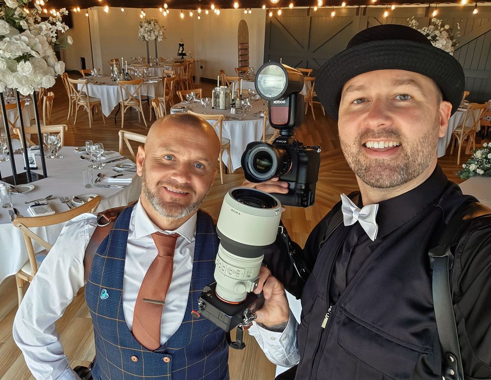 Two men smile for a selfie at a wedding venue, one holding a professional camera with a large lens and flash.