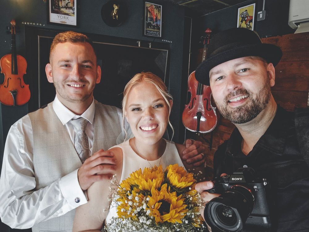 Wedding photographer poses with couple holding sunflower bouquet in music-themed venue.