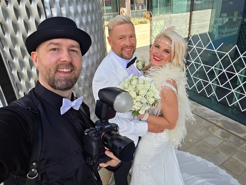 Professional wedding photographer takes selfie with newlyweds holding bouquet outside modern building.