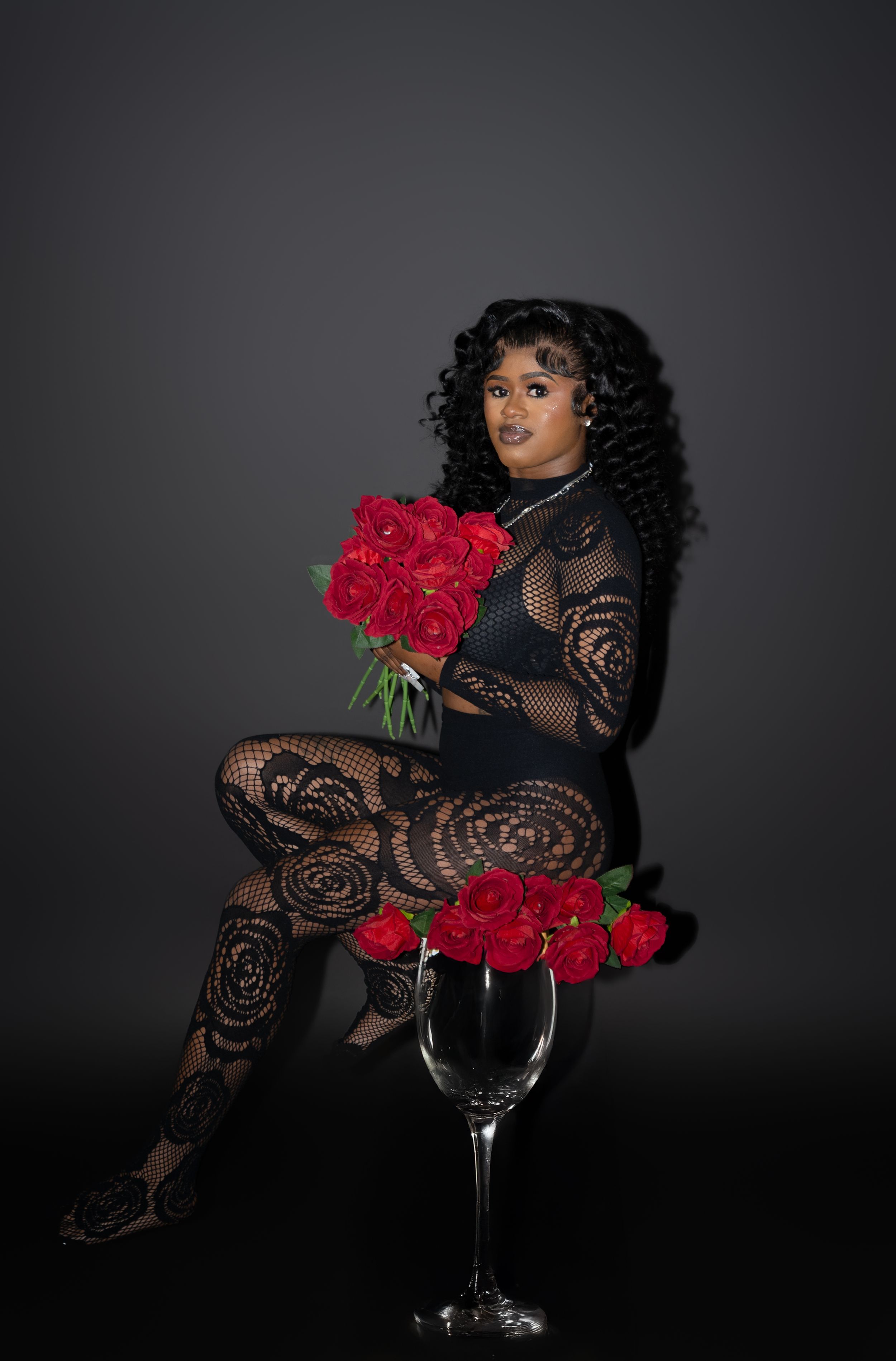 A woman in a black lace bodysuit sits gracefully holding red roses, with a large wine glass filled with roses beside her.