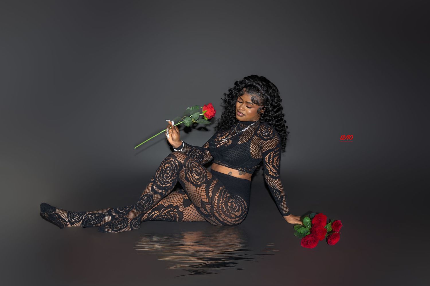 Woman in black lace outfit poses with red roses on reflective surface against gray background.