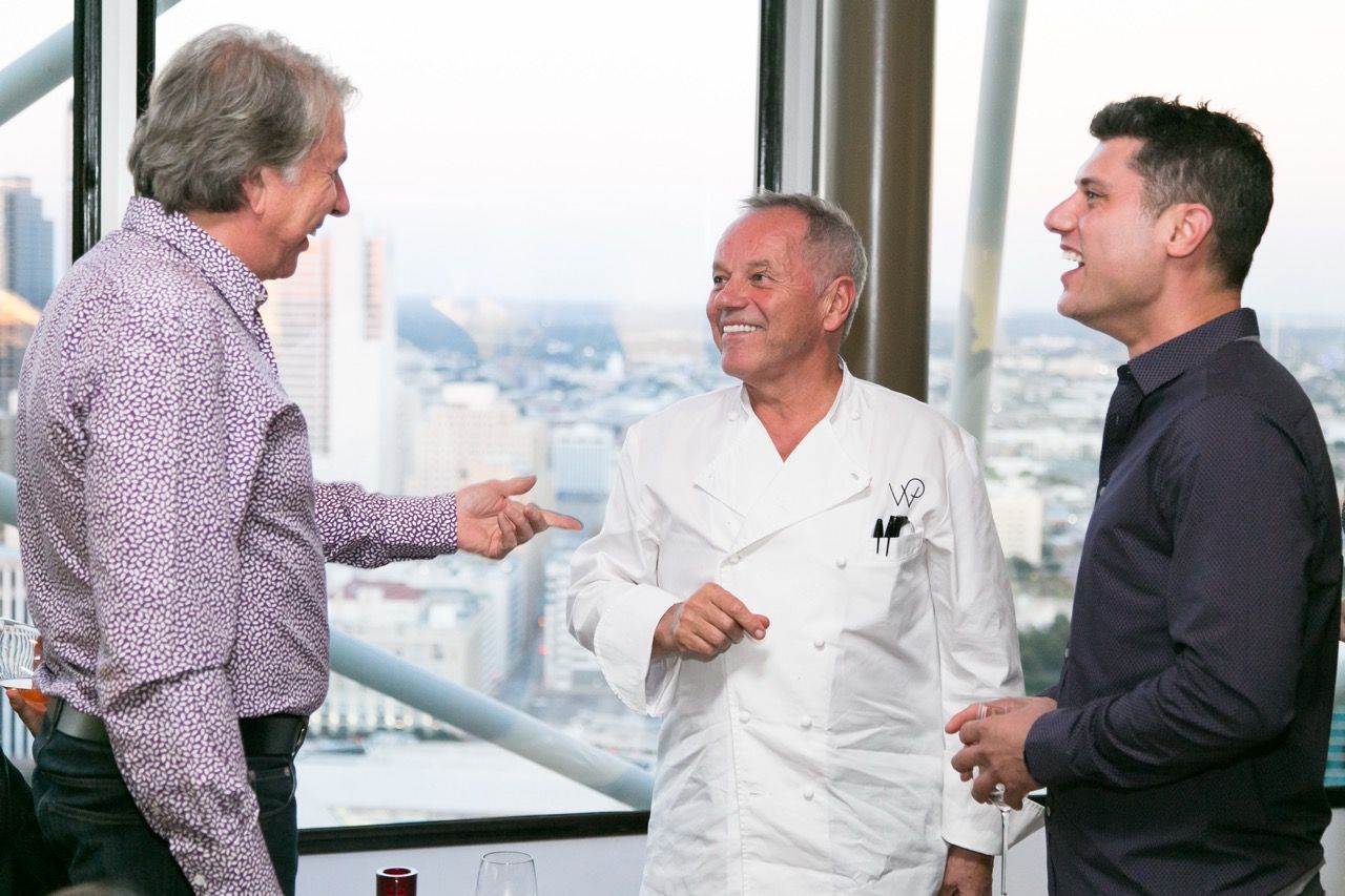 Celebrity chef Wolfgang Puck speaking with colleagues near a window in Reunion Tower with the Dallas city skyline view behind them