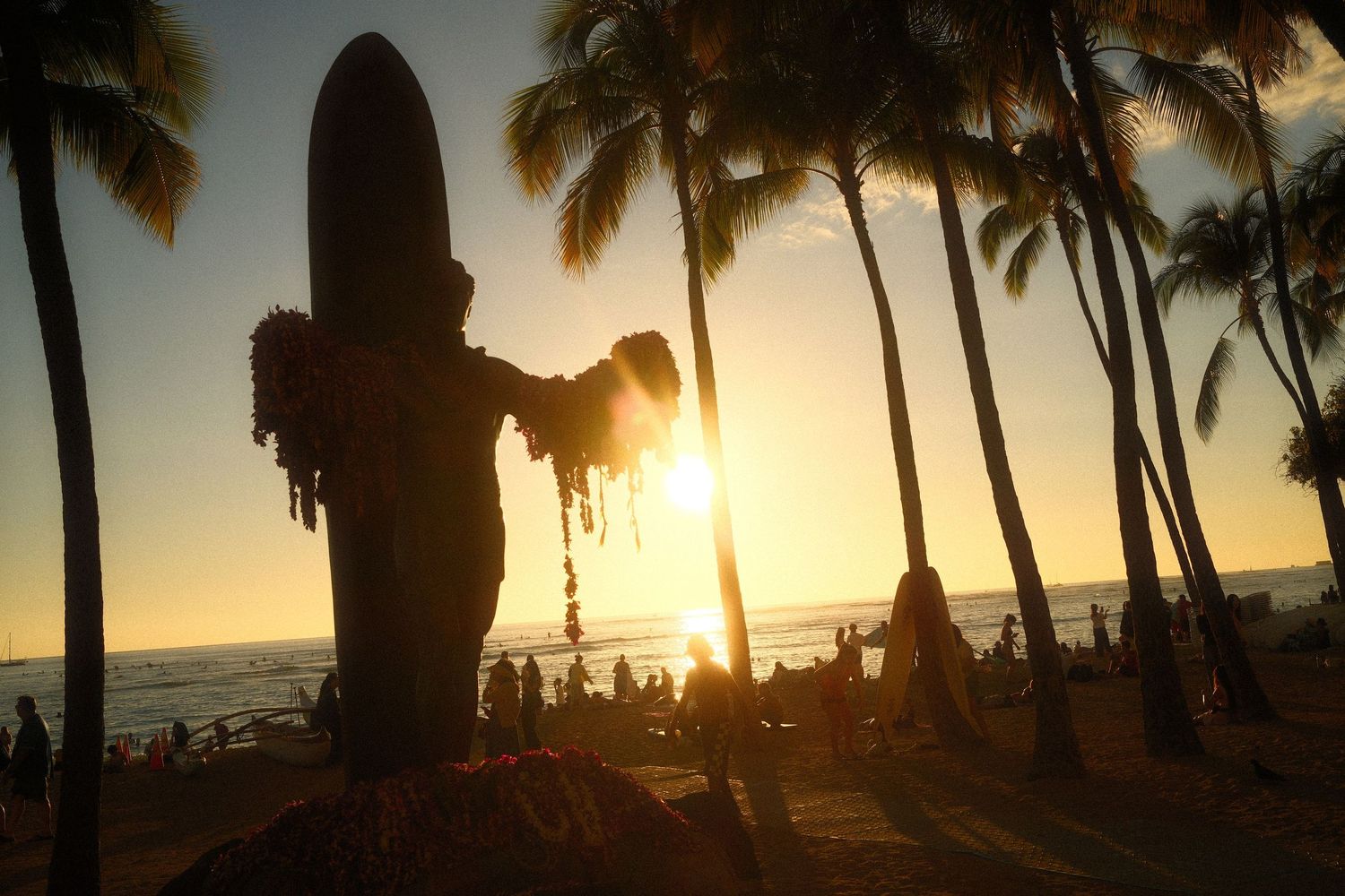 Silhouette of a surfer statue at sunset on Waikiki Beach with palm trees and ocean in the background.