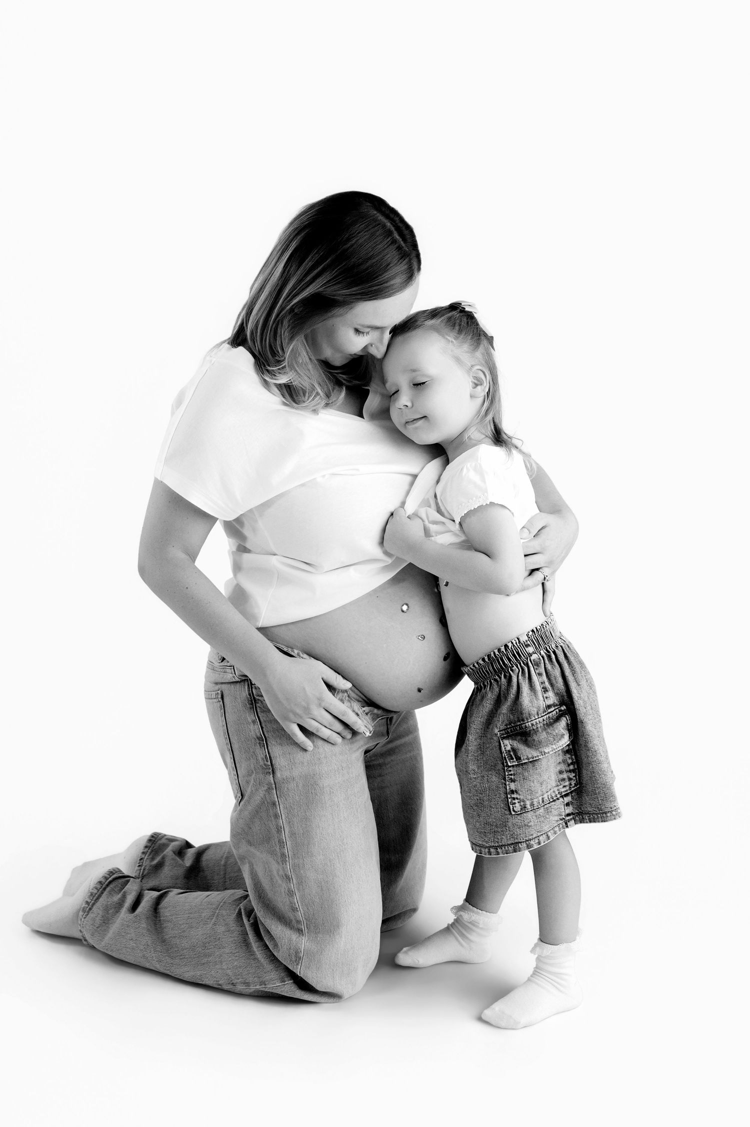 Pregnant woman in flowing white fabric poses gracefully against white background, cradling her baby bump.