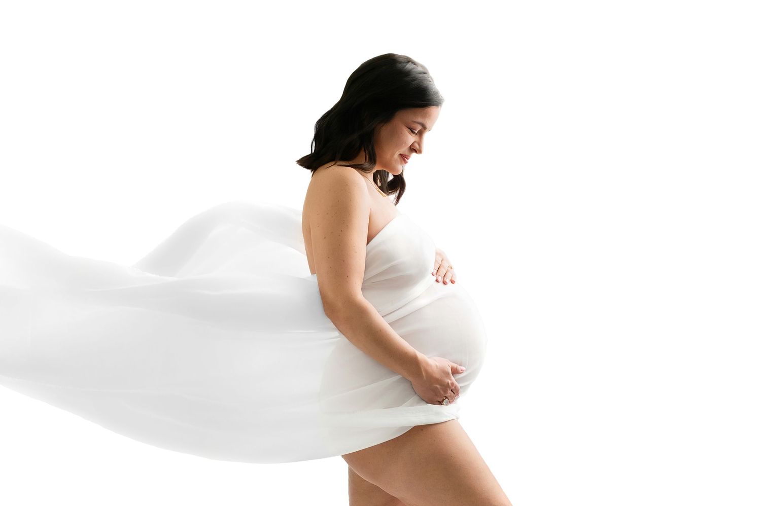 Pregnant woman in flowing white fabric poses gracefully against white background, cradling her baby bump.