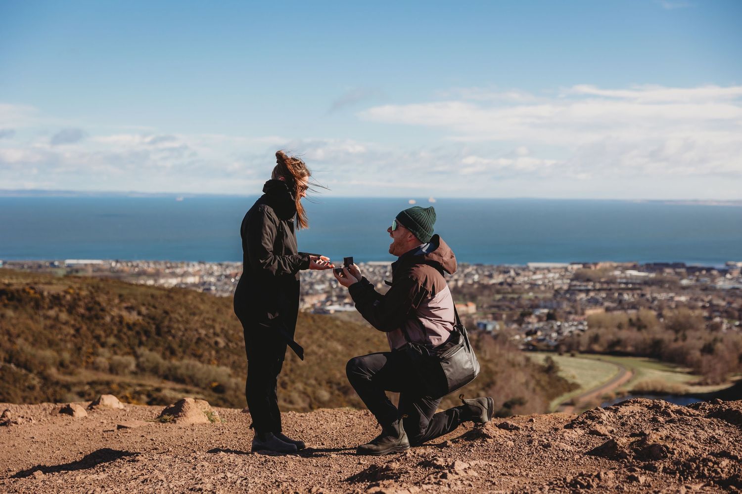 Two hikers stand on a rocky hilltop overlooking a coastal city under a bright blue sky.