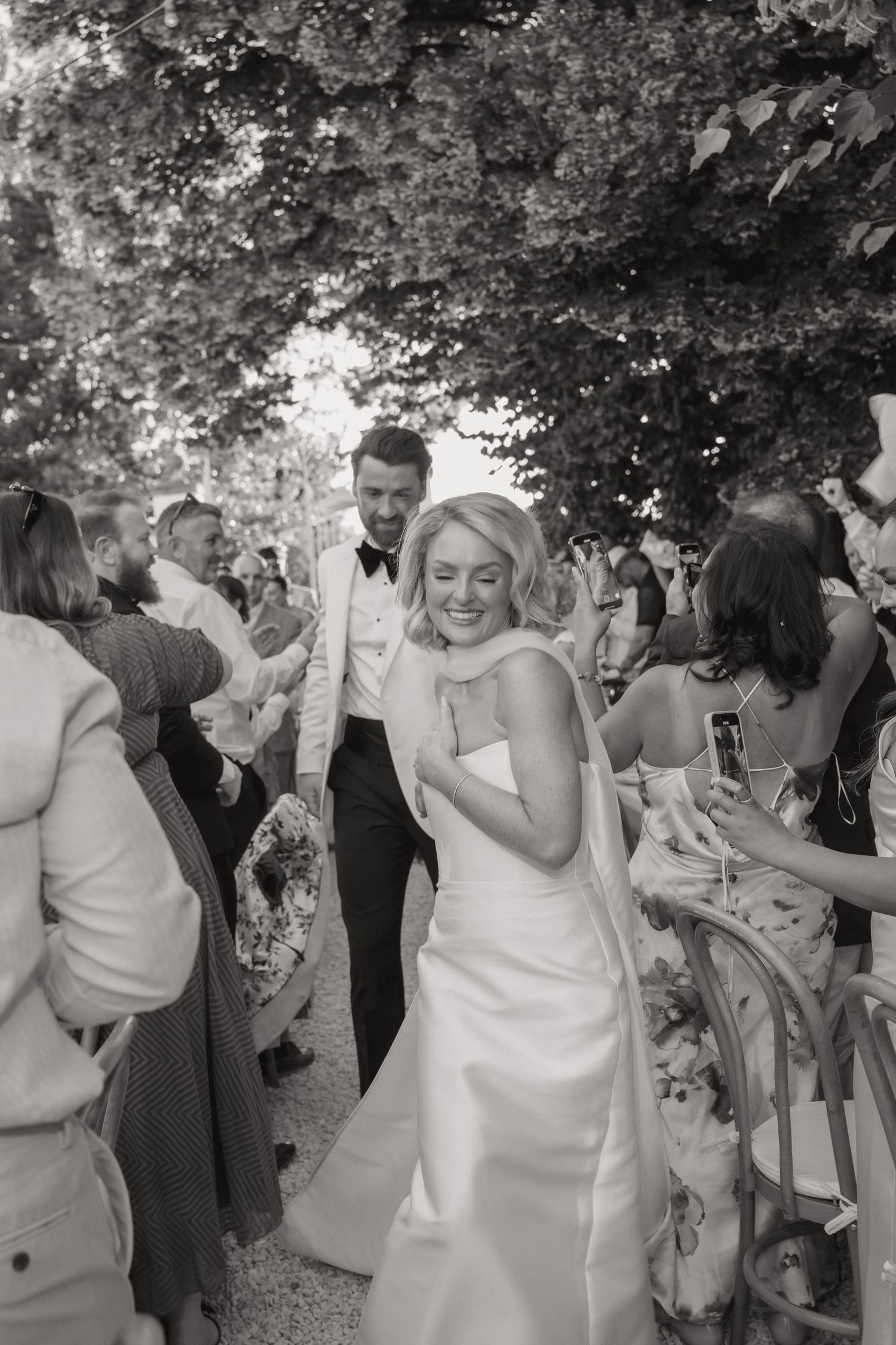 A joyful wedding celebration outdoors under trees with guests and a bride in a white strapless gown dancing and mingling.