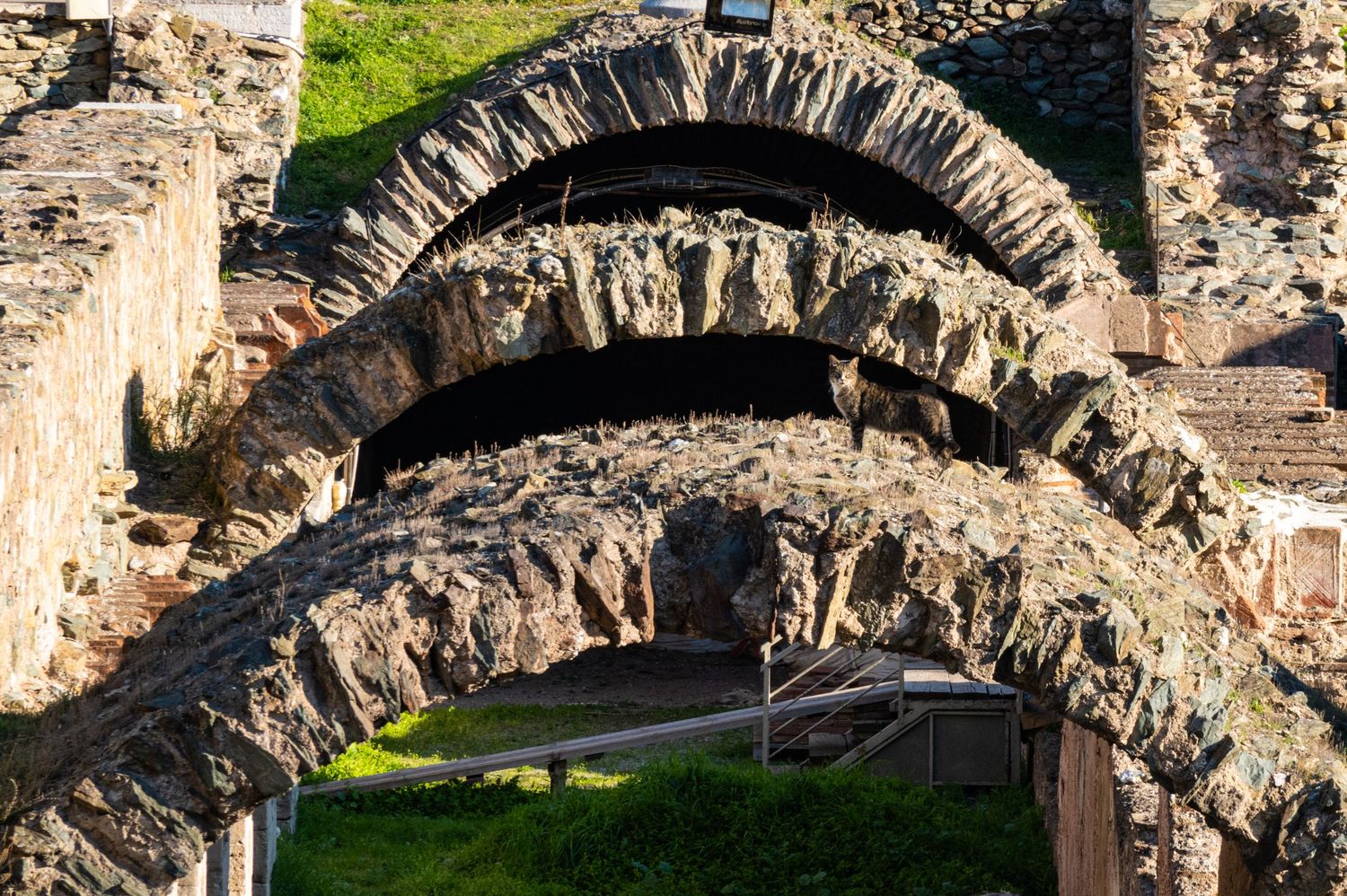 Ancient stone archways and ruins of a historic fortress wall stretch across a green landscape in daylight.
