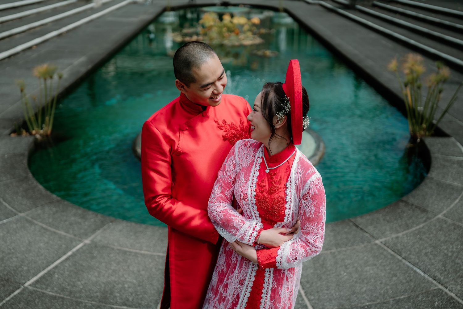 A couple in traditional red and pink wedding attire stands together by a turquoise fountain in a garden setting.