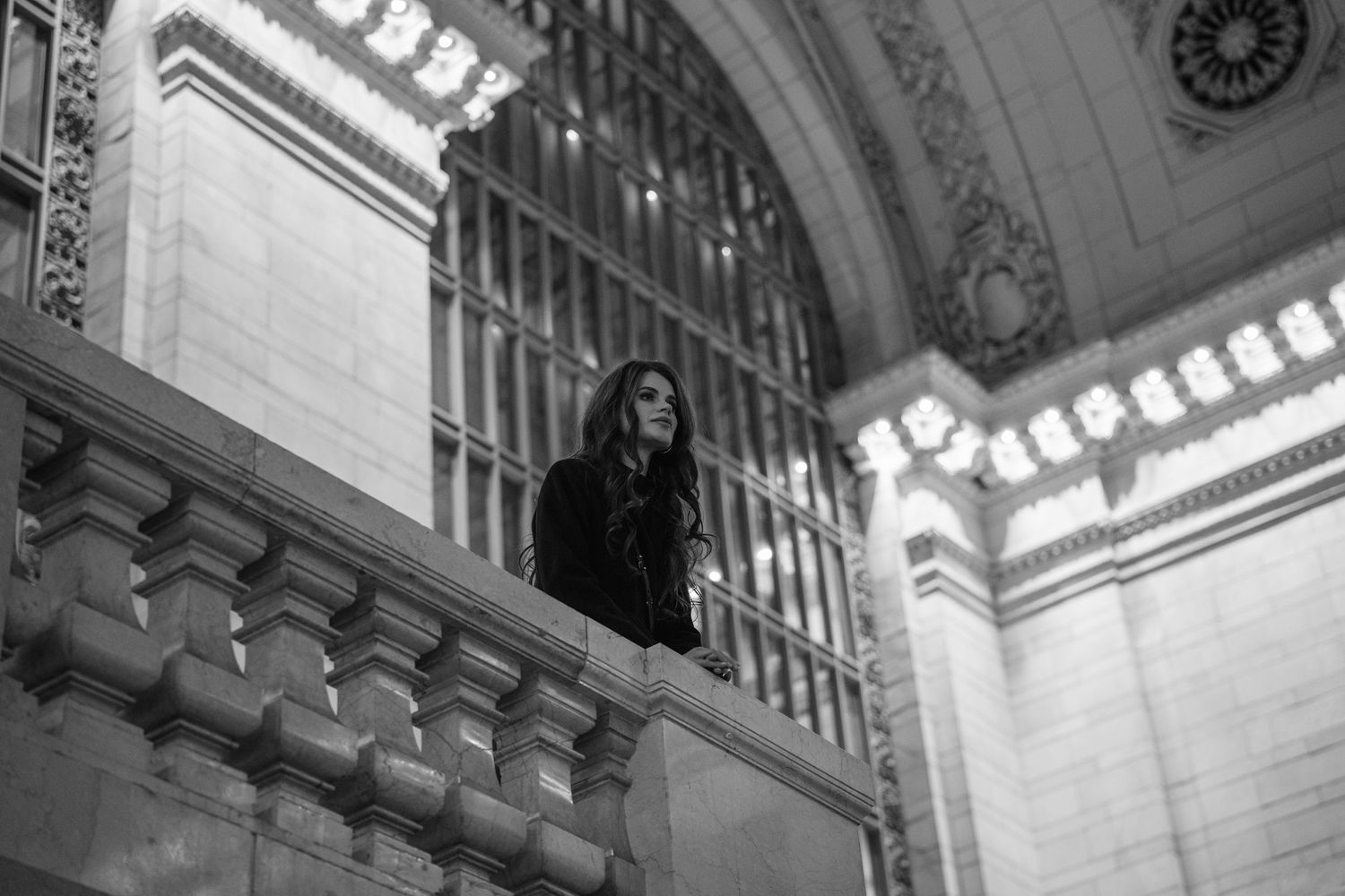 A young woman stands on a balcony in a grand historic building in black and white photography.