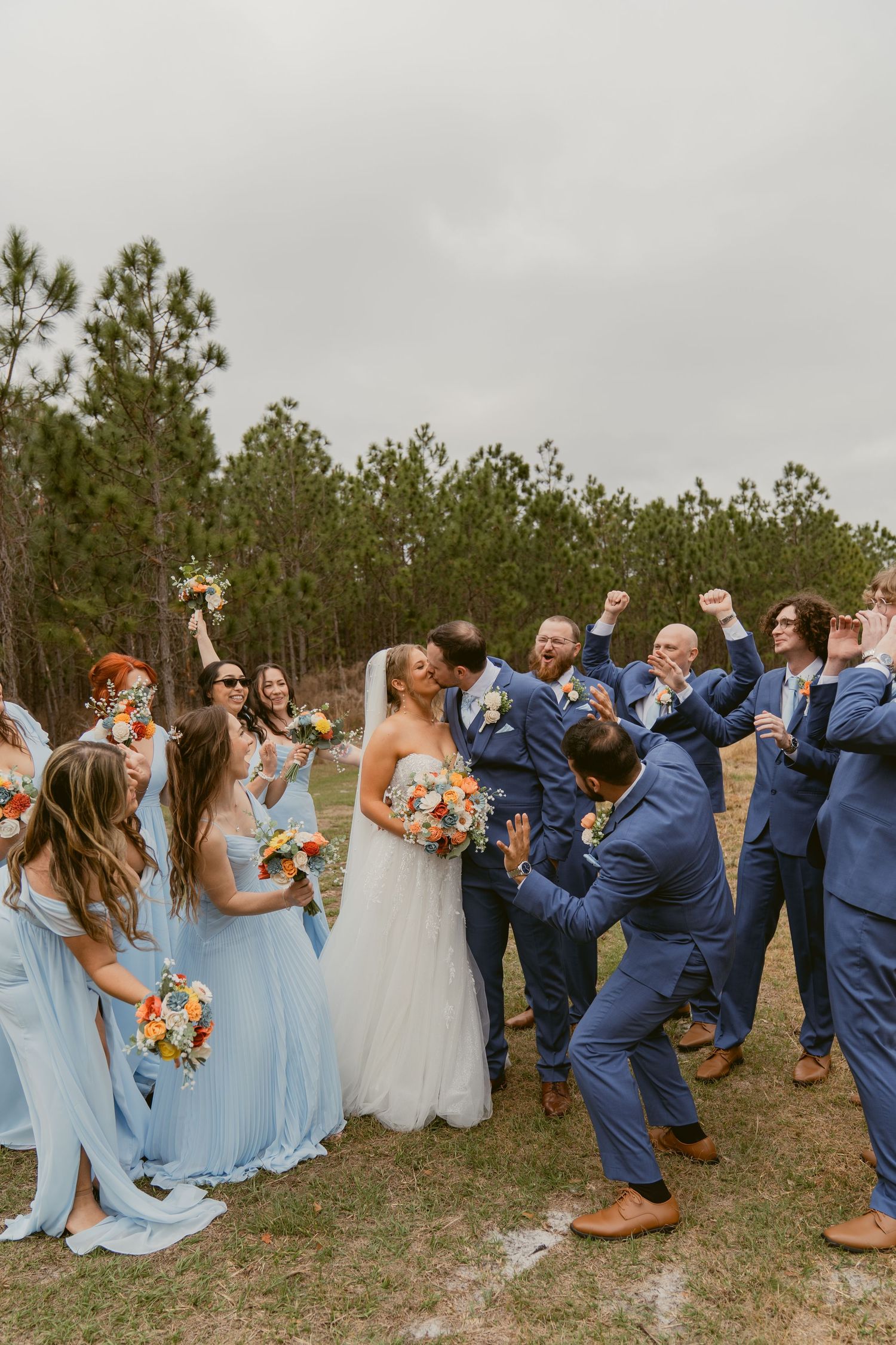 Wedding party members walk and laugh together in an outdoor field setting wearing formal attire and pastel bridesmaid dresses.