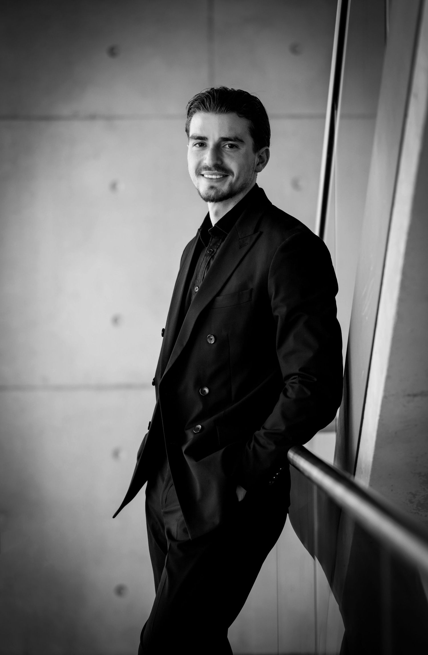 Schwarz-Weiß-Businessportrait eines Mannes am Treppengeländer, lächelnd mit den Händen in den Hosentaschen.