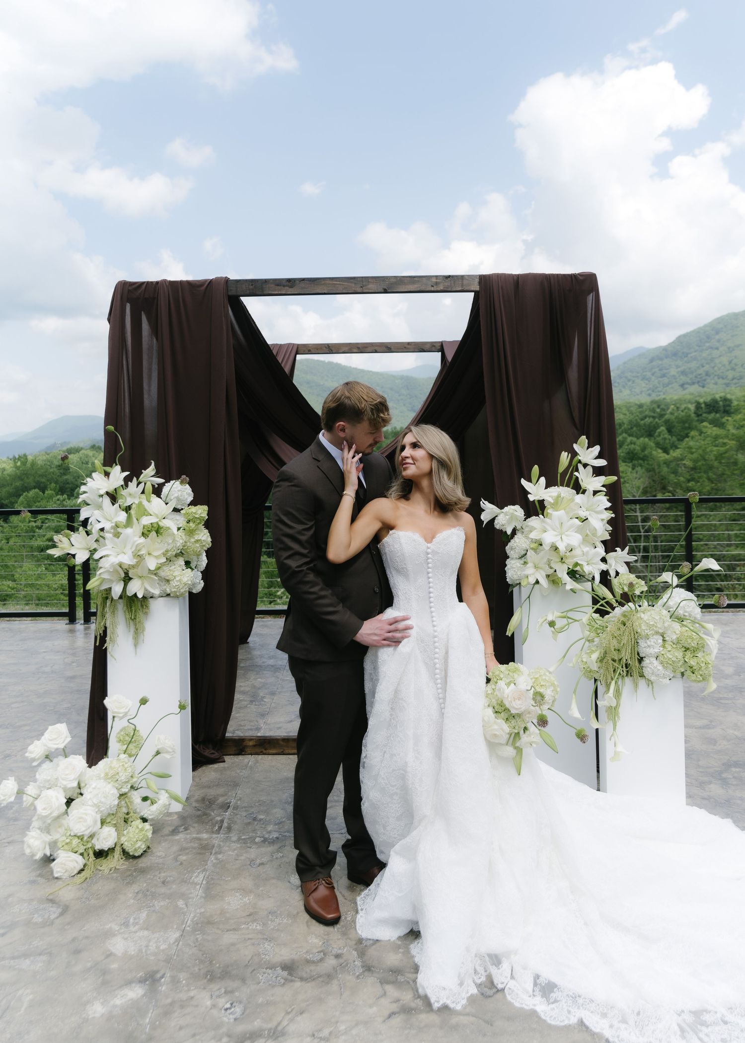 Bride and groom share a romantic moment under a dark-draped arch with white floral arrangements and mountain views.