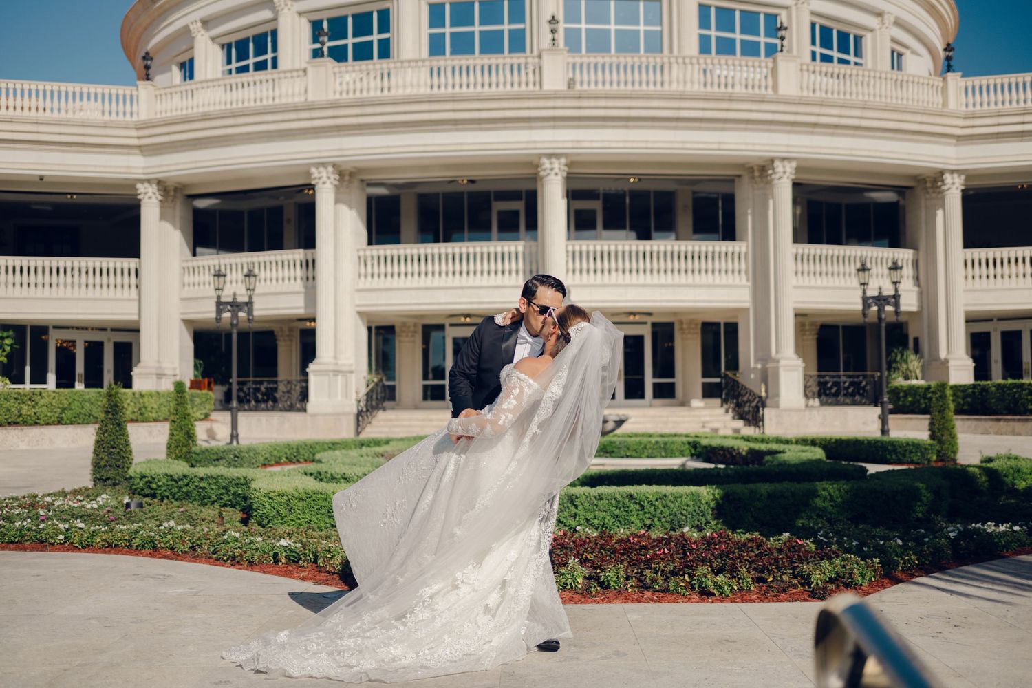 A bride and groom share a romantic moment outside an elegant white columned building with curved architecture.