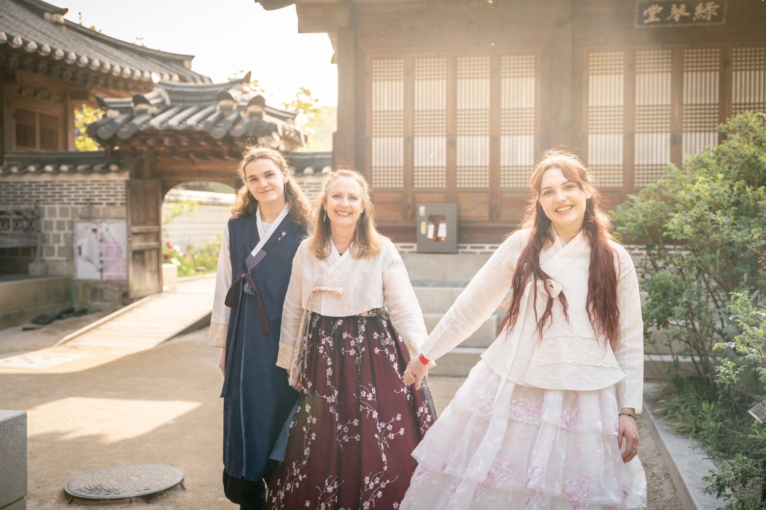 Friends in traditional Korean hanbok dresses pose together at a historic temple courtyard in warm sunlight.