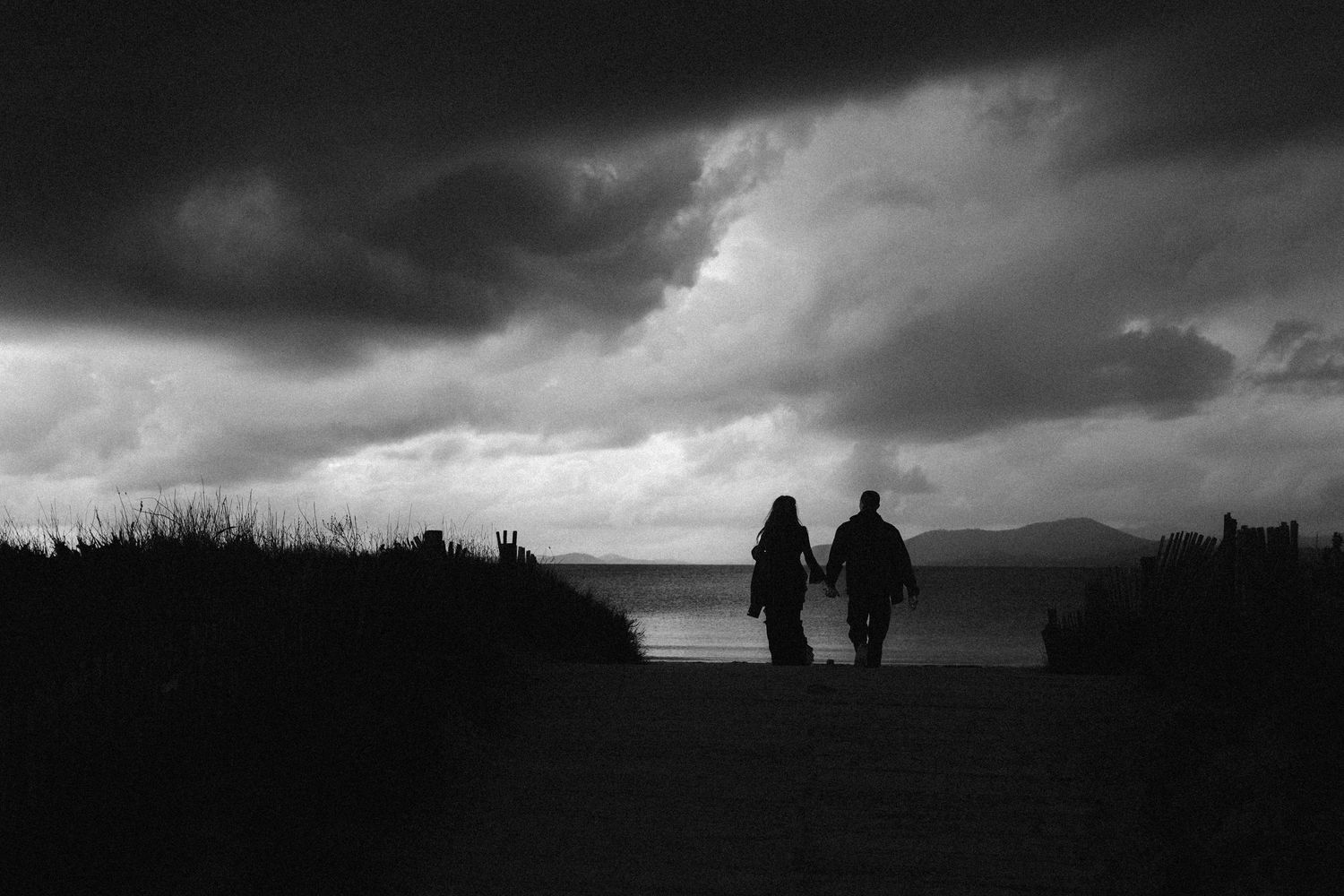 Silhouetted figures stand against dramatic stormy skies in a series of moody black and white landscape photographs.