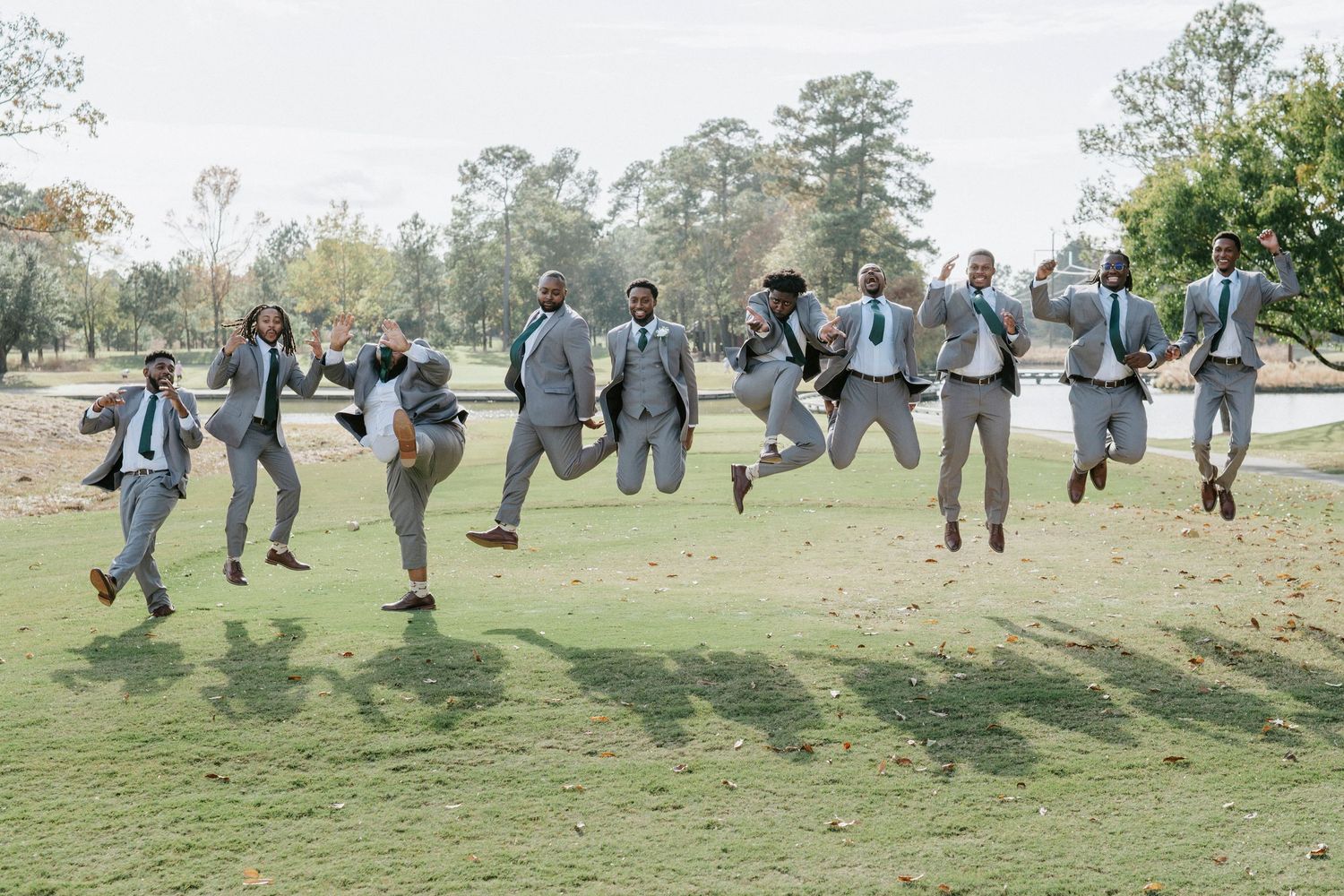 Wedding party in light gray suits leaping joyfully together on a grassy lawn during outdoor celebration.
