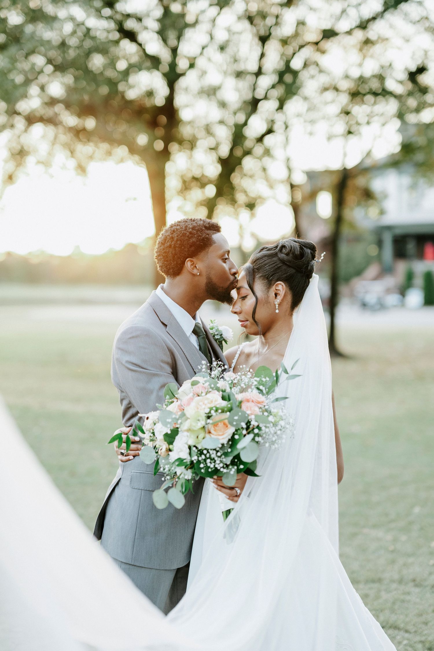 Romantic outdoor wedding portraits show couple embracing in a sunlit park setting with white floral bouquet.