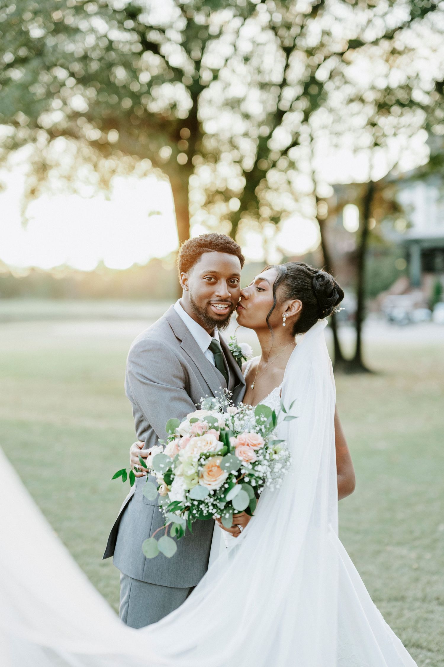 A romantic wedding couple embraces outdoors in a park setting with soft lighting and floral bouquet details.