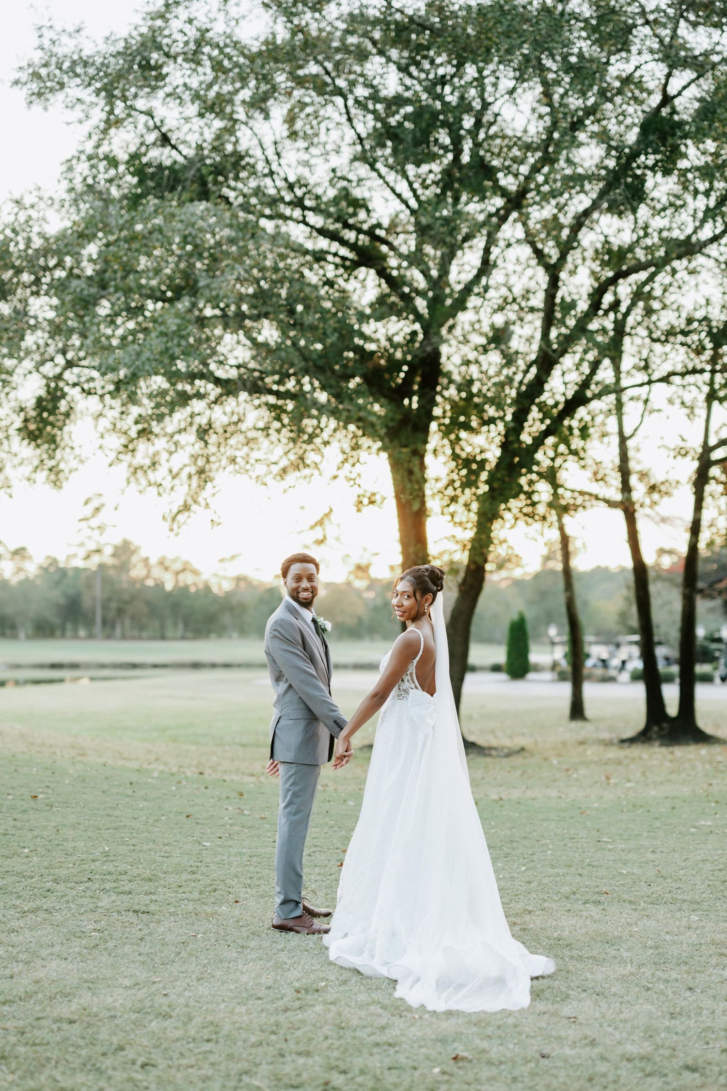 A bride and groom share a joyful moment dancing together in a sunlit field with trees at golden hour.