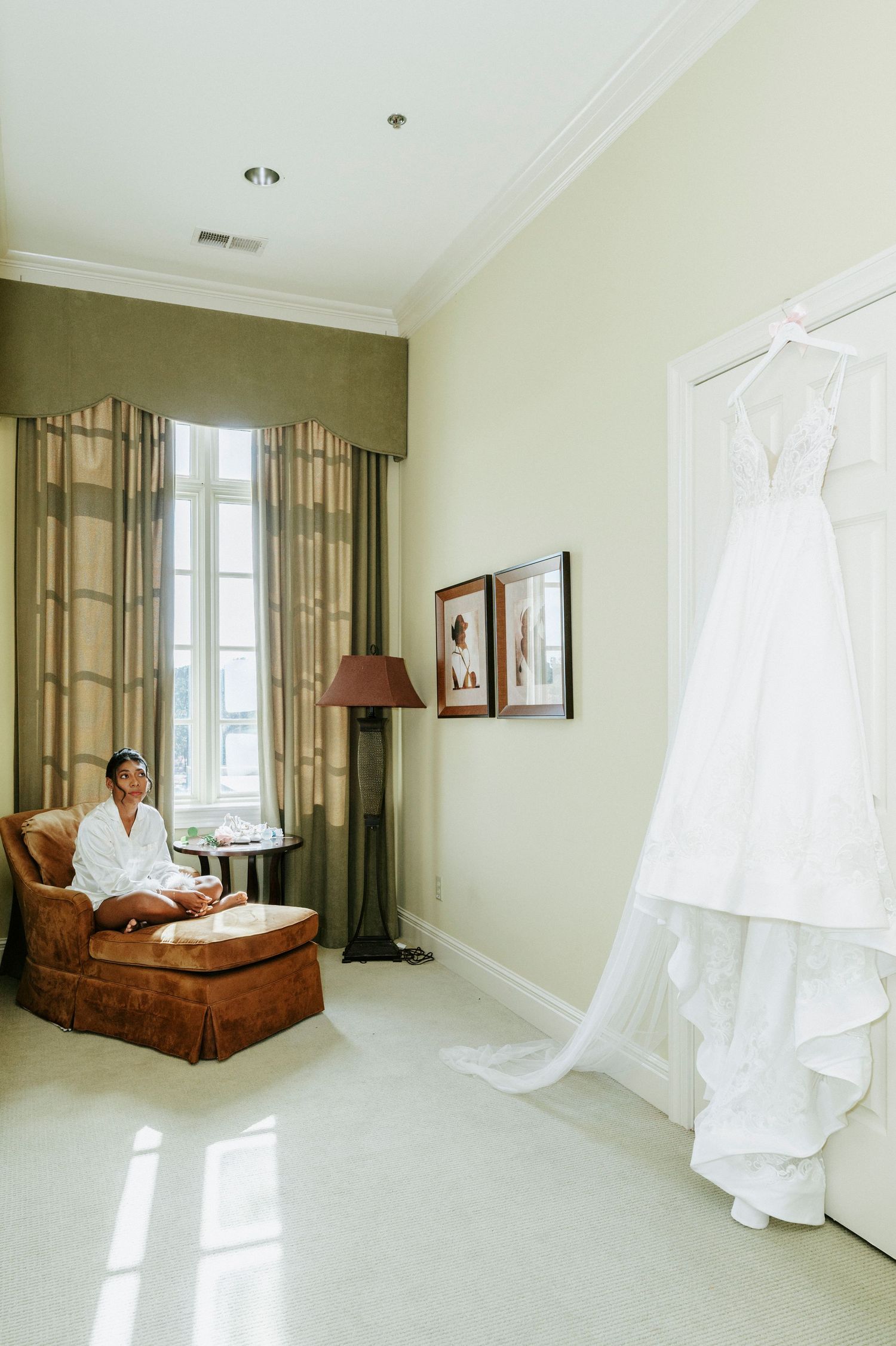 Elegant hotel room interior with white wedding dress displayed and brown chaise lounge against neutral walls.