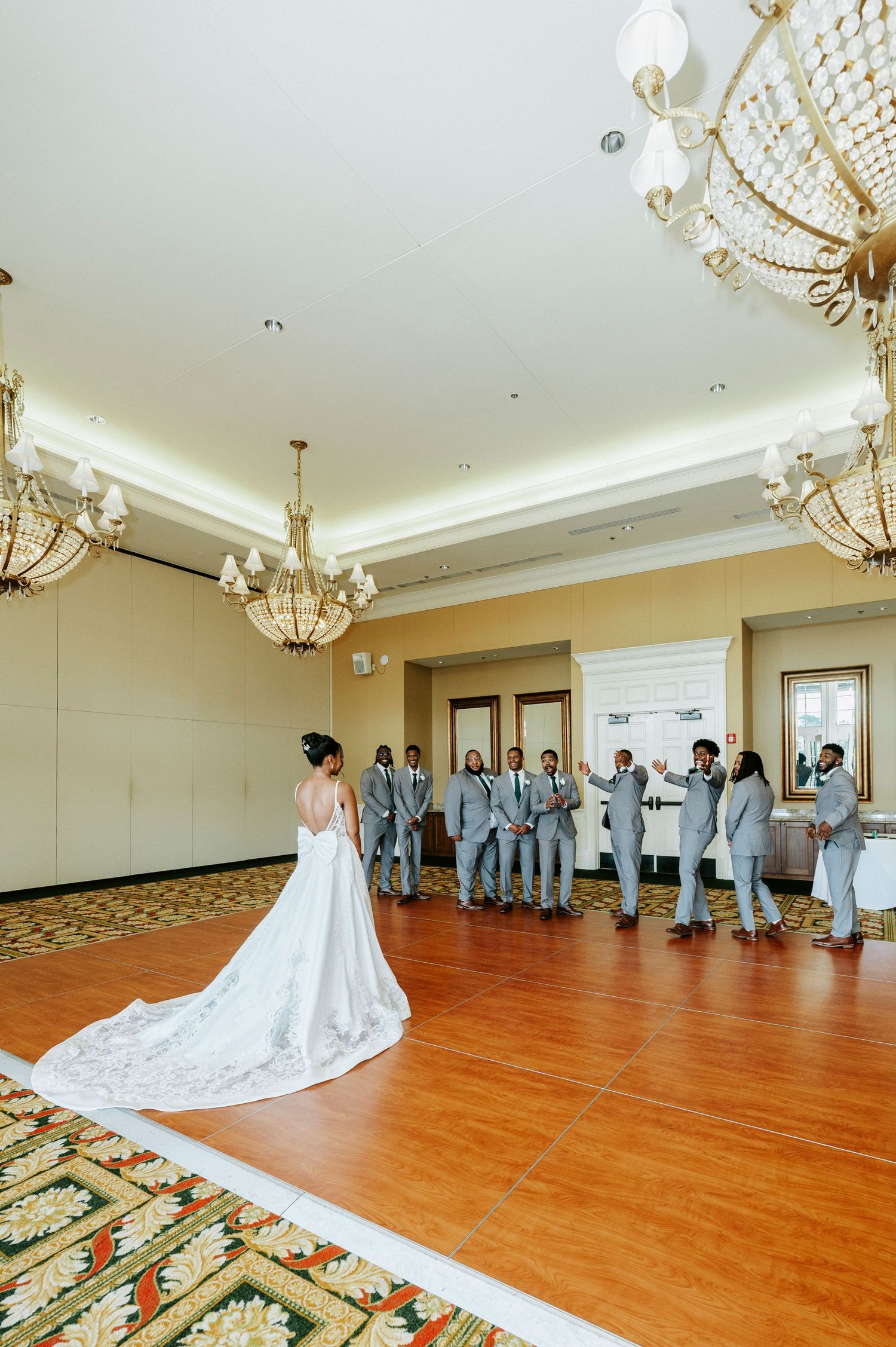 A wedding party gathers in an elegant ballroom with chandeliers and hardwood floors during a formal ceremony.