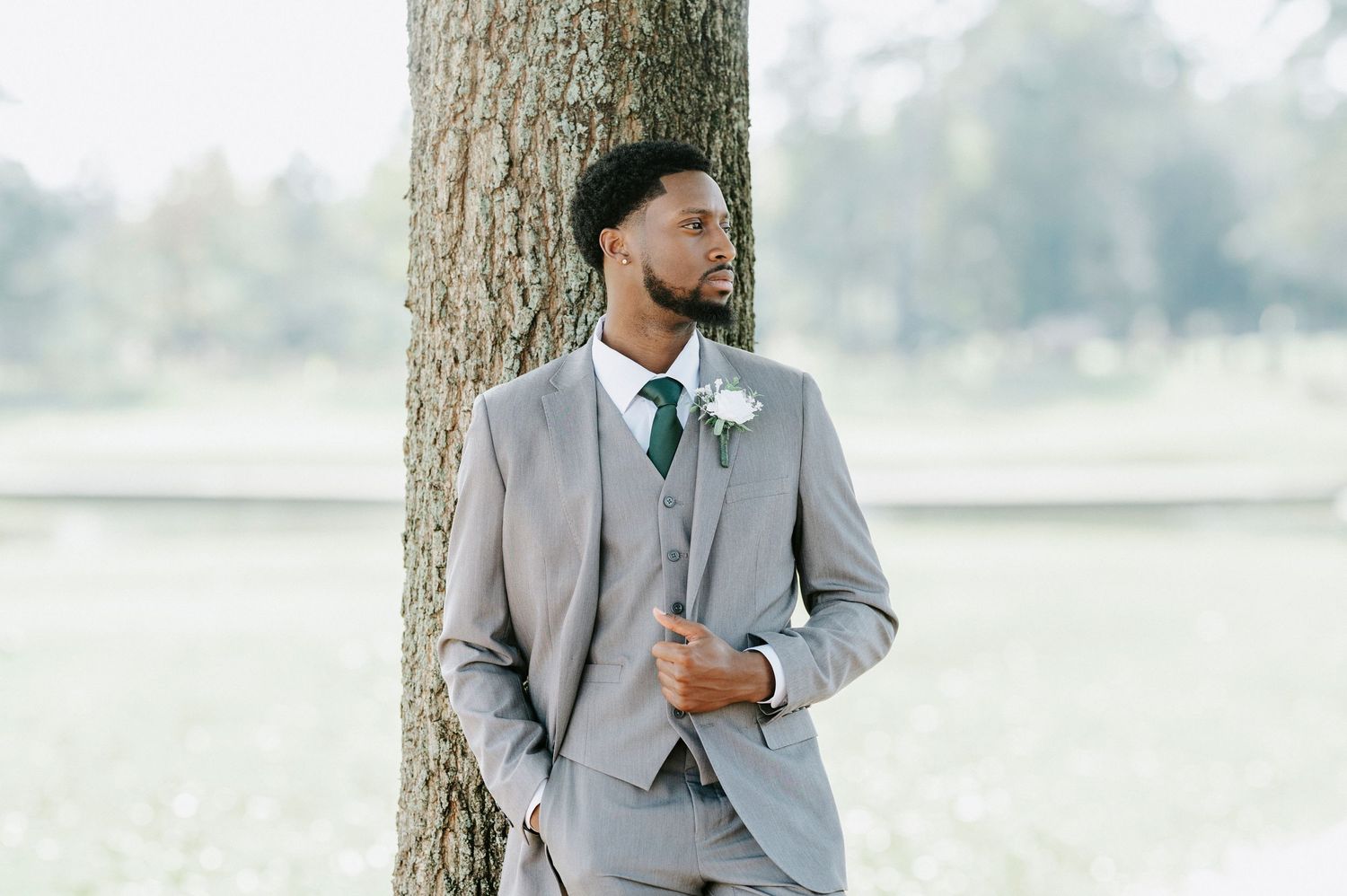 A groom in a light gray suit and green tie poses casually against a tree trunk in an outdoor setting.