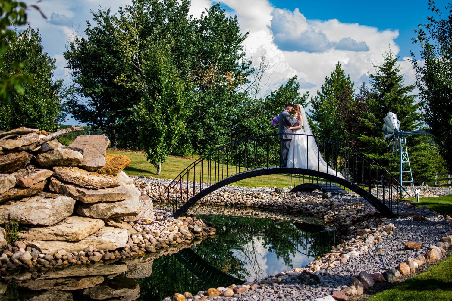 A tranquil waterfall cascades over stone ledges in a landscaped garden with evergreen trees and blue sky.