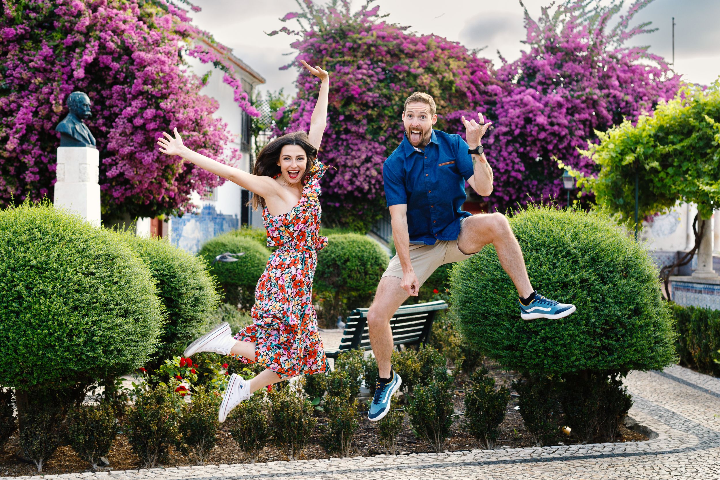 Playful couple jumping and posing in front of vibrant purple bougainvillea flowers in a garden setting.