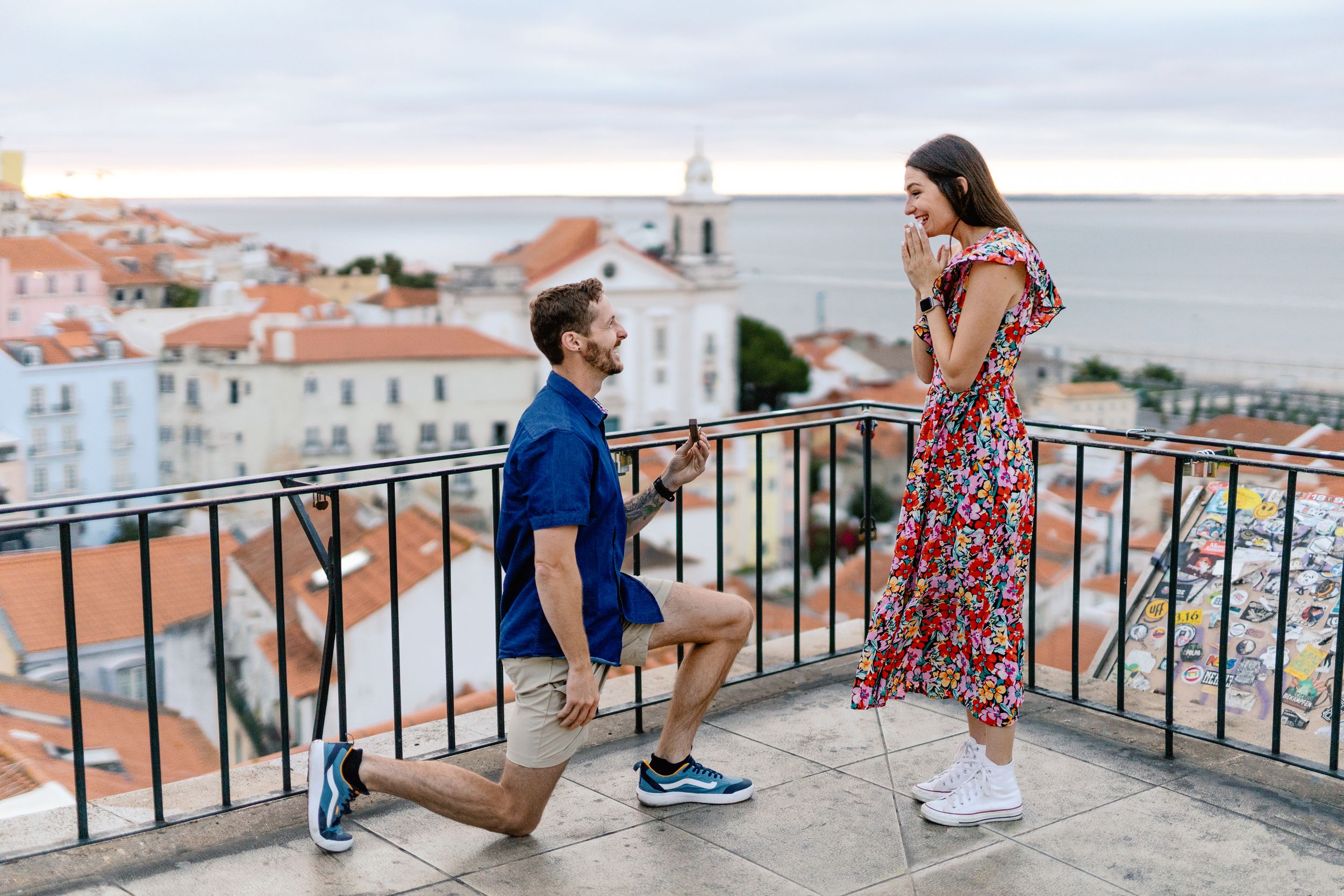 A couple shares a romantic moment on a balcony overlooking a coastal Mediterranean town with terracotta rooftops.