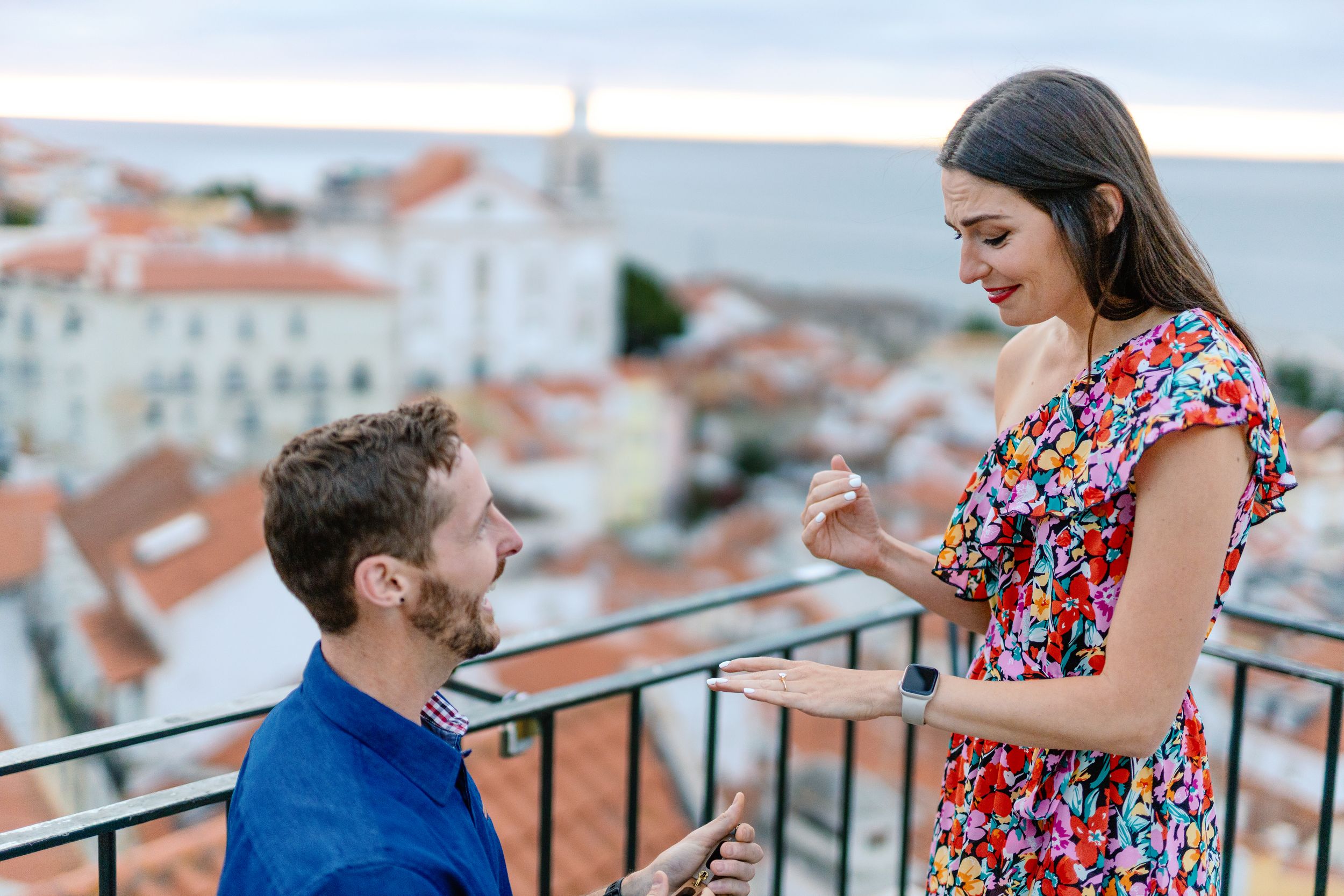 A couple shares a romantic moment on a balcony overlooking a European cityscape with red-tiled roofs.