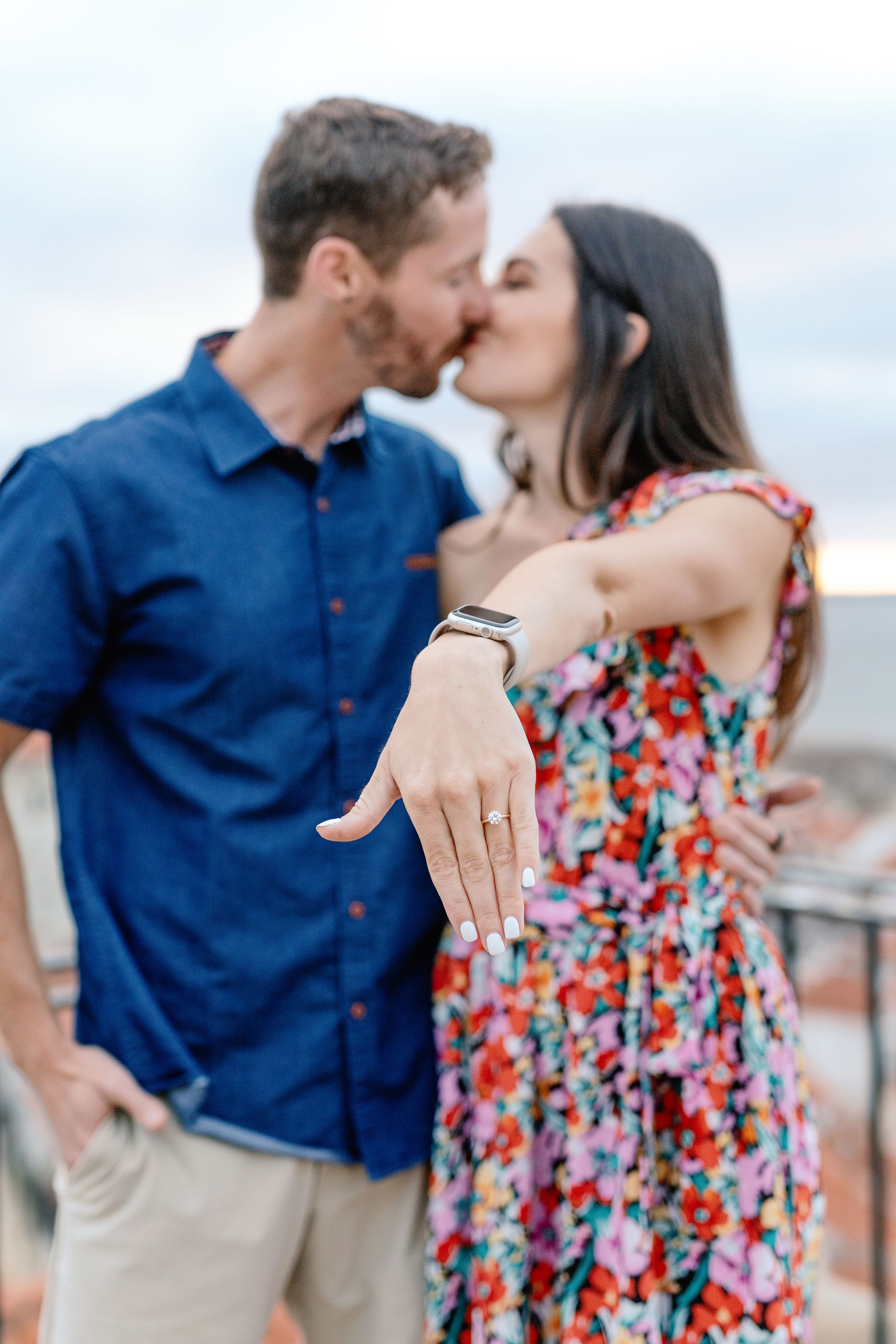An engaged couple shares a romantic moment on a terrace while showing off a new engagement ring.