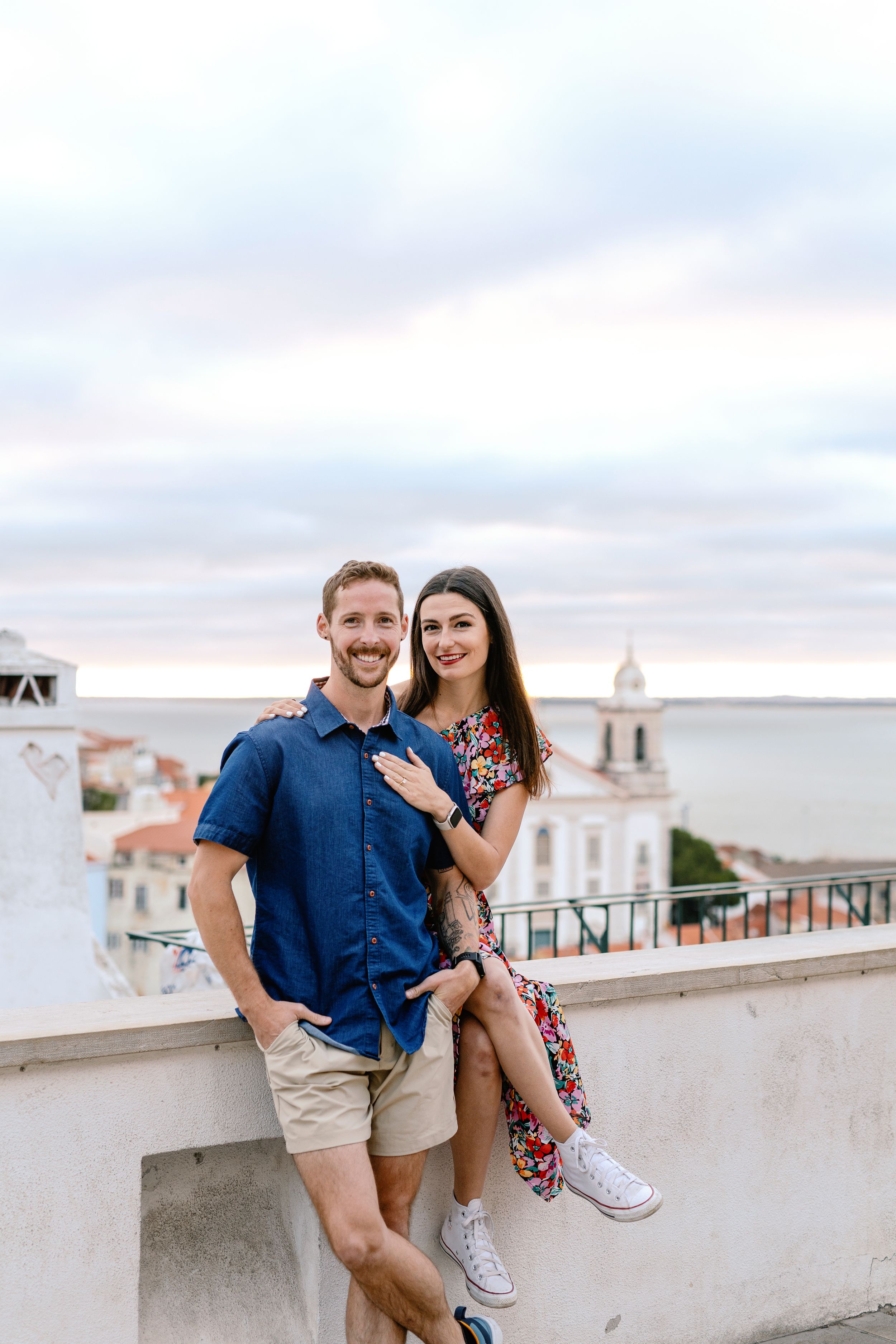 A couple poses together on a rooftop terrace overlooking a Mediterranean coastal town with white buildings and church spires.