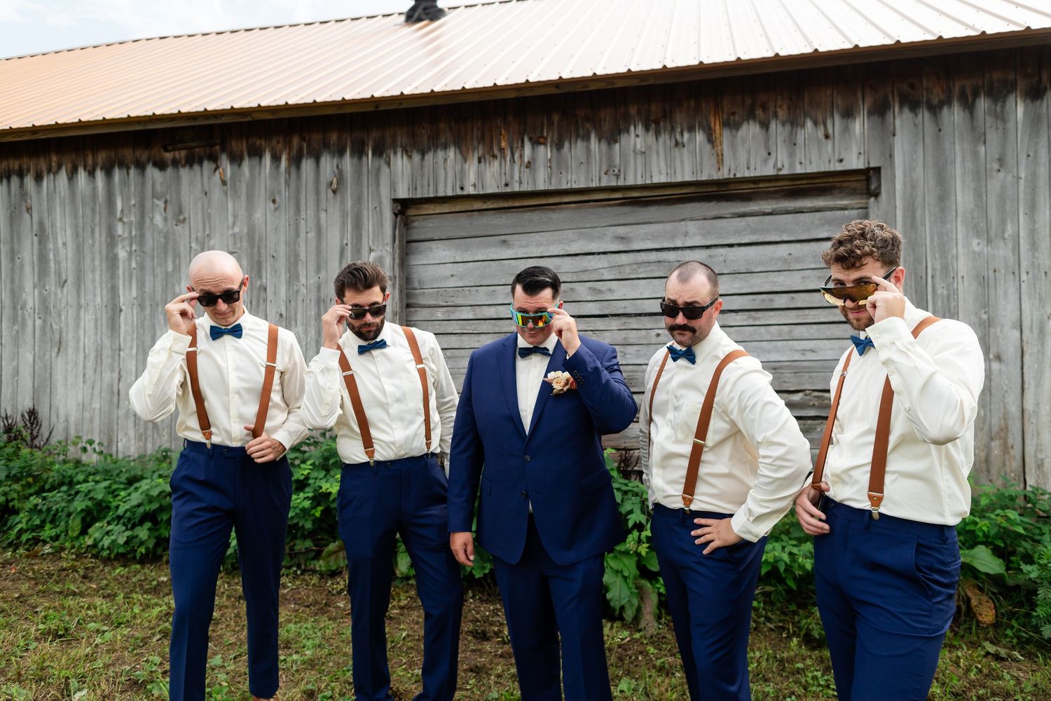 Groomsmen in navy pants and suspenders share a drink together while standing in front of rustic barn wall. Minnesota Wisconsin Wedding Photo