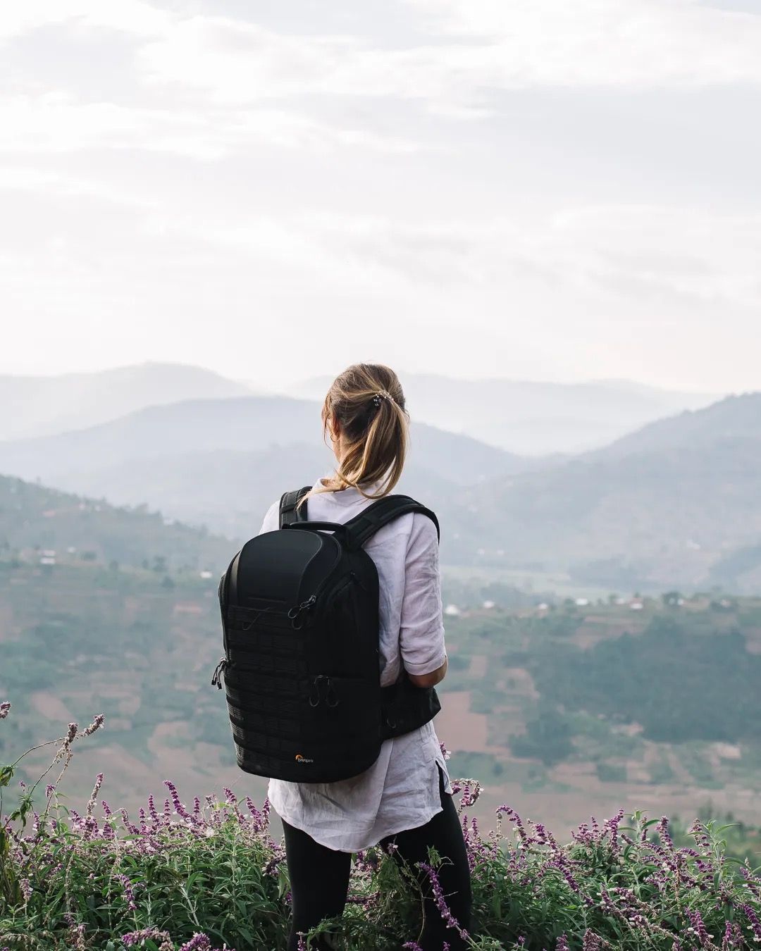Woman with backpack stands on hillside overlooking a misty mountain valley, capturing adventure and peaceful nature views.