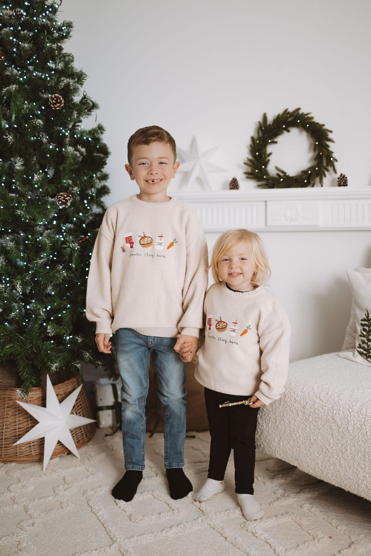 Two children wearing matching cream sweaters stand in front of a Christmas tree and white mantel with star decorations.