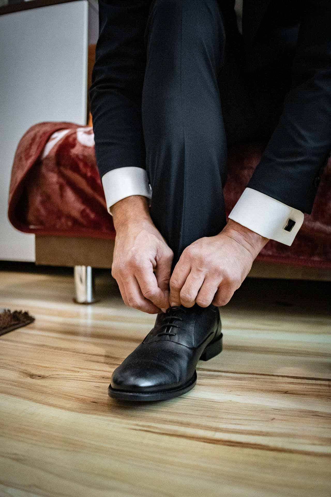 Man putting on black formal dress shoes on wooden floor.