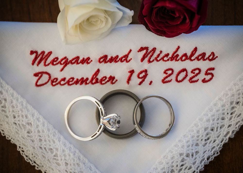 Wedding rings displayed on white lace with red and white roses and names and date text overlay.