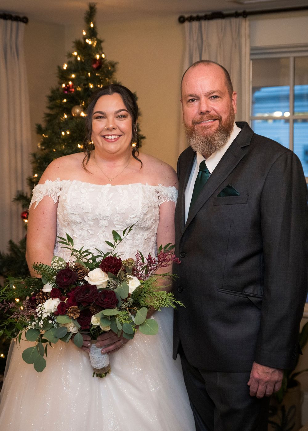 Wedding couple poses in formal attire with burgundy and white floral bouquet near a decorated Christmas tree.