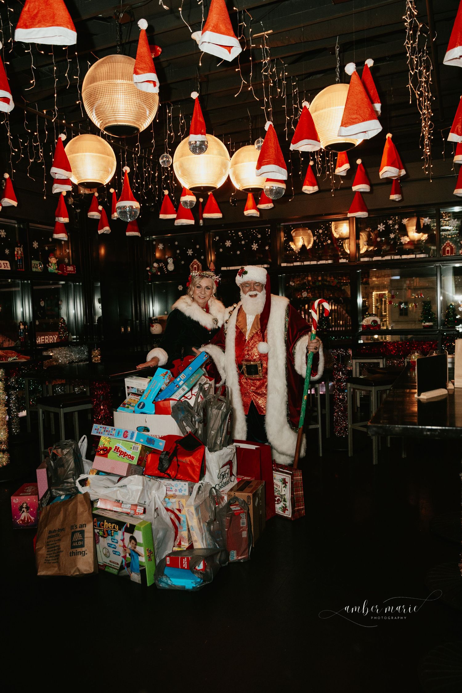 Christmas decorations with Santa hats adorn glowing pendant lights above a large pile of wrapped holiday presents in a dark room.