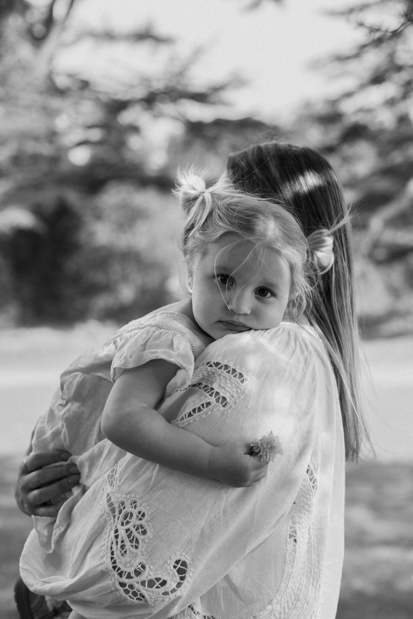 Black and white photo of a woman cuddling a young girl who is holding a flower in Brighton woodland