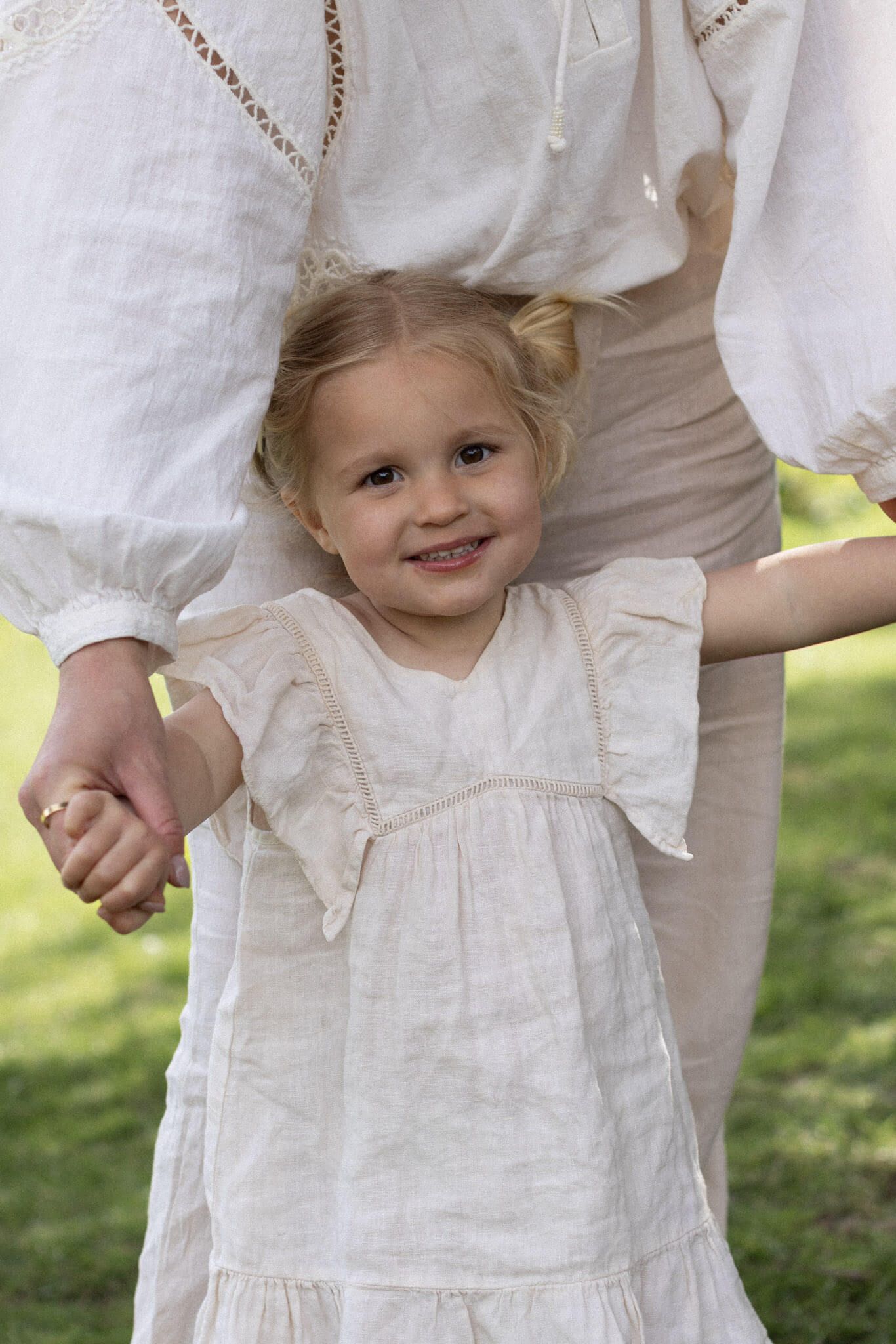 A smiling toddler girl in a neutral ruffled dress holds her mother's hand during an outdoor photography session in a Brighton park