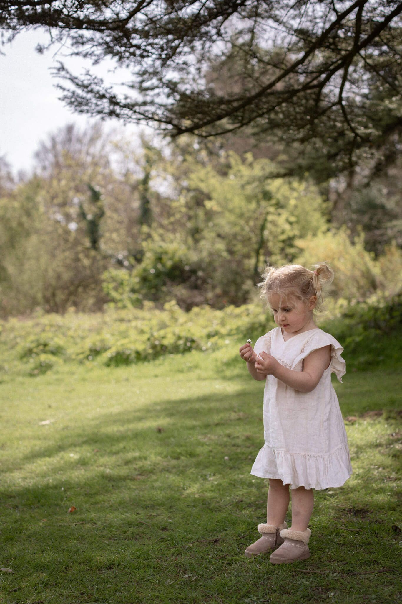 Toddler girl photographed in Brighton woodland dressed in a neutral dress and holding and looking at a flower
