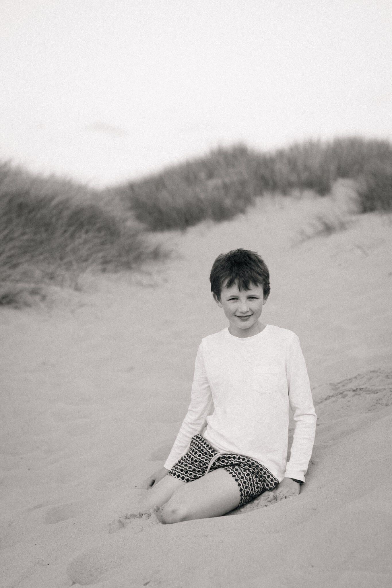 A black and white photo of a smiling boy sitting on sandy beach dunes during an outdoor family photography shoot