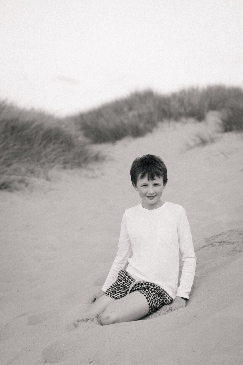 A black and white photo of a smiling boy sitting on sandy beach dunes during an outdoor family photography shoot