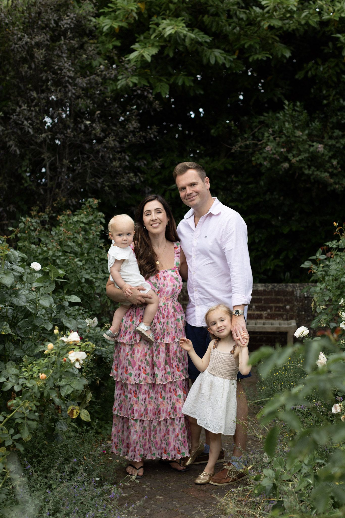 Family of four posing in a lush garden during a family photo shoot, mother in floral dress holding baby, father with young daughter.