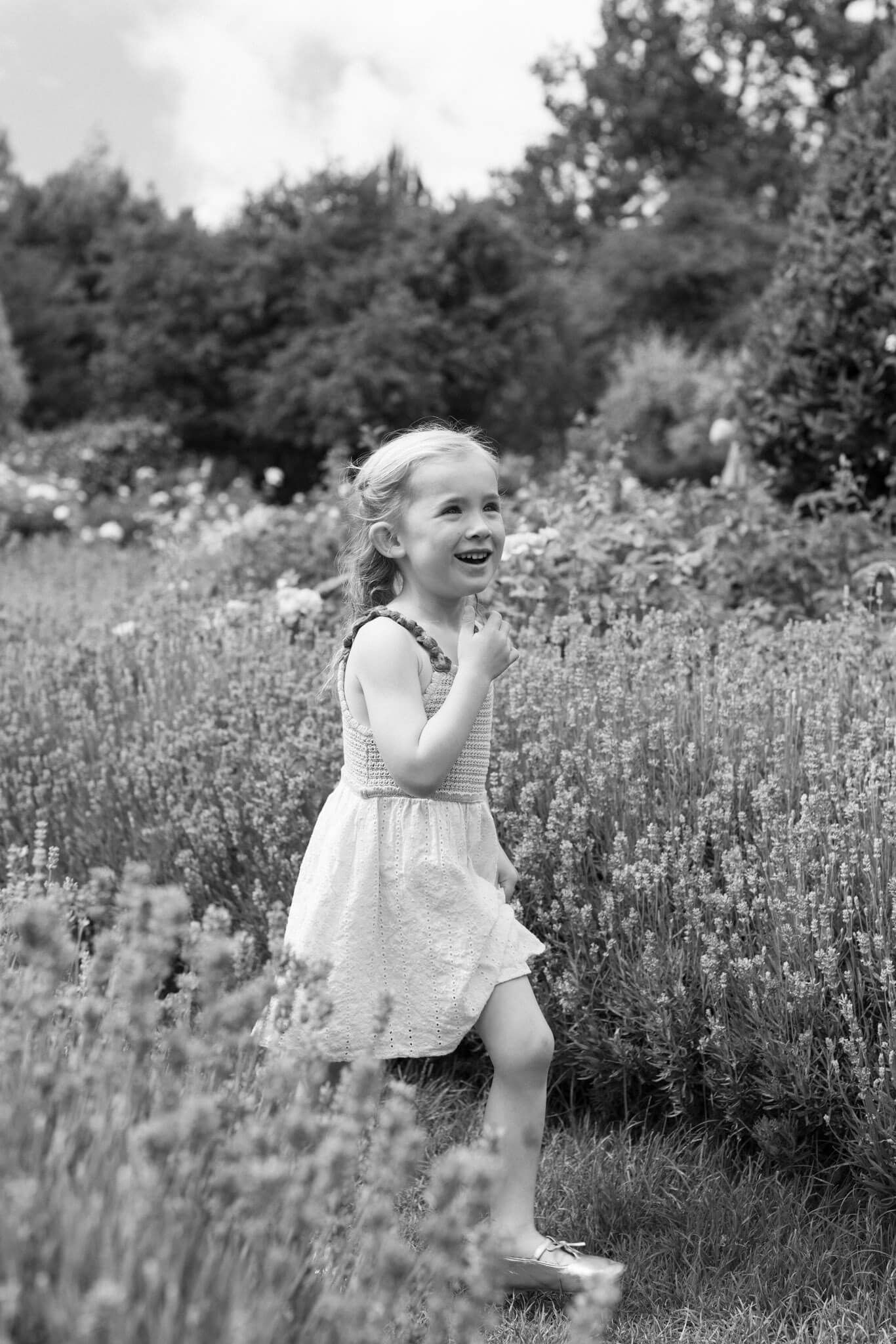 Black and white photo of a joyful young girl in a white dress laughing in a blooming Brighton garden with lavender flowers.