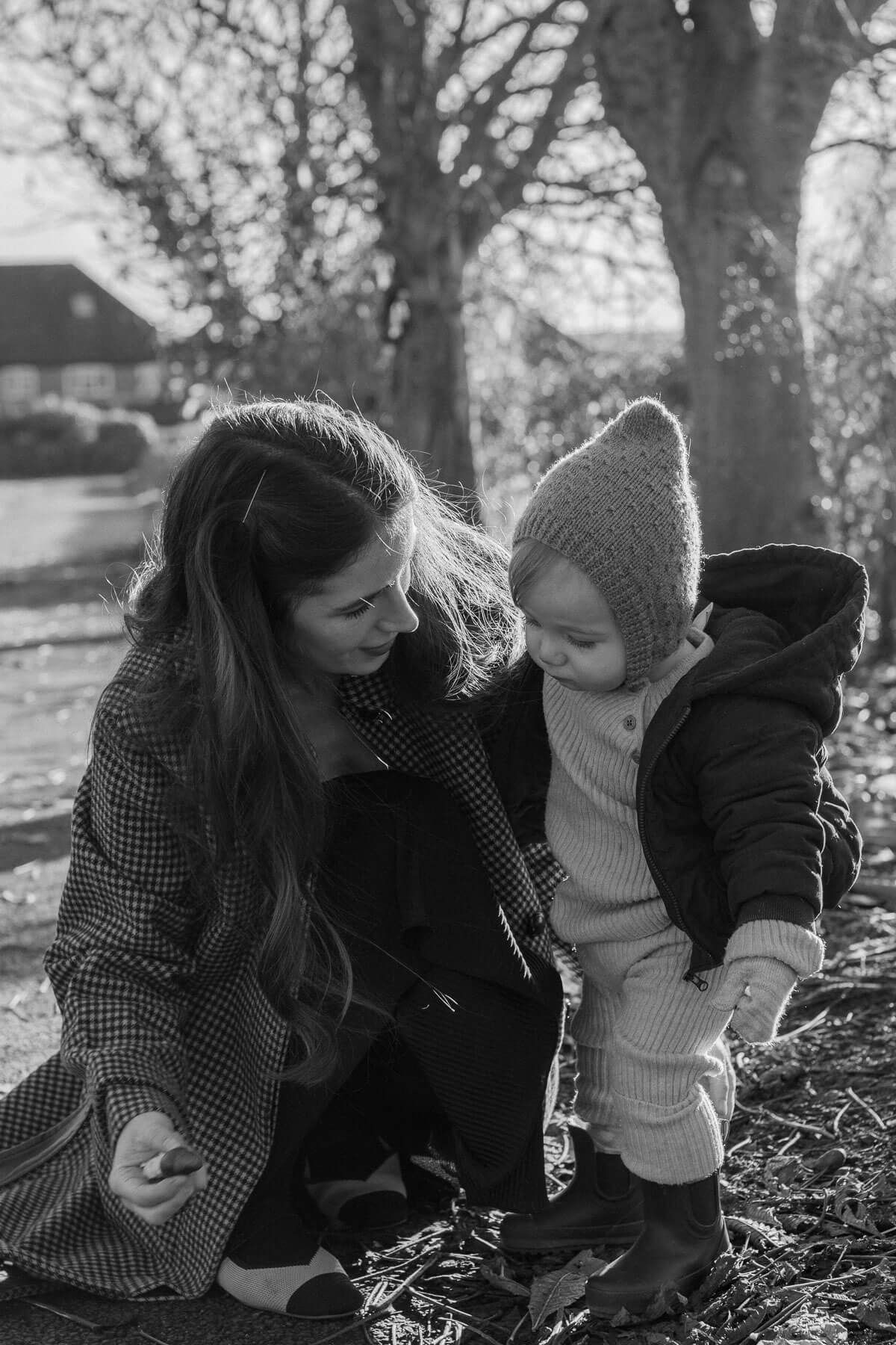 Black and white photo of mother crouching down looking at a leaf with her child, photographed in natural light in Sussex woodland