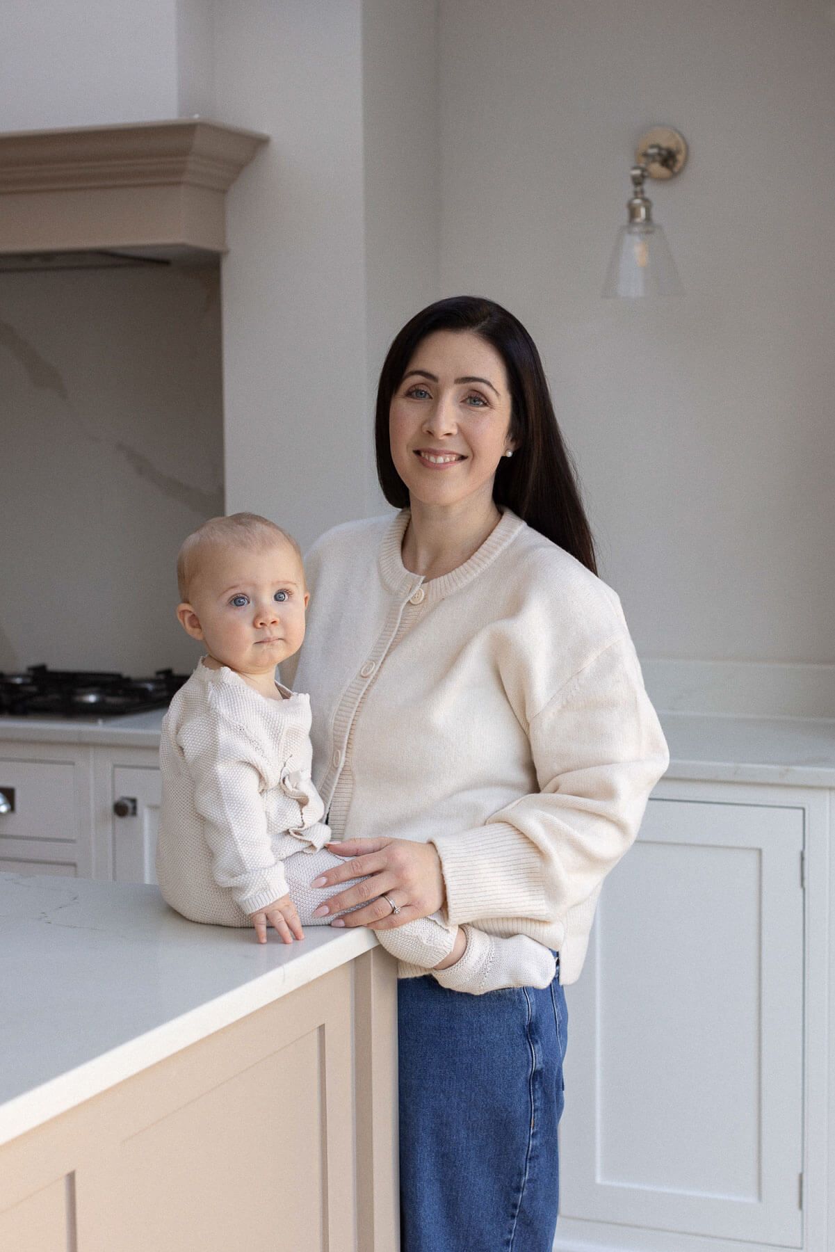 Elegant portrait of mother and baby standing in a bright, naturally lit kitchen, photographed during an in home session in Brighton