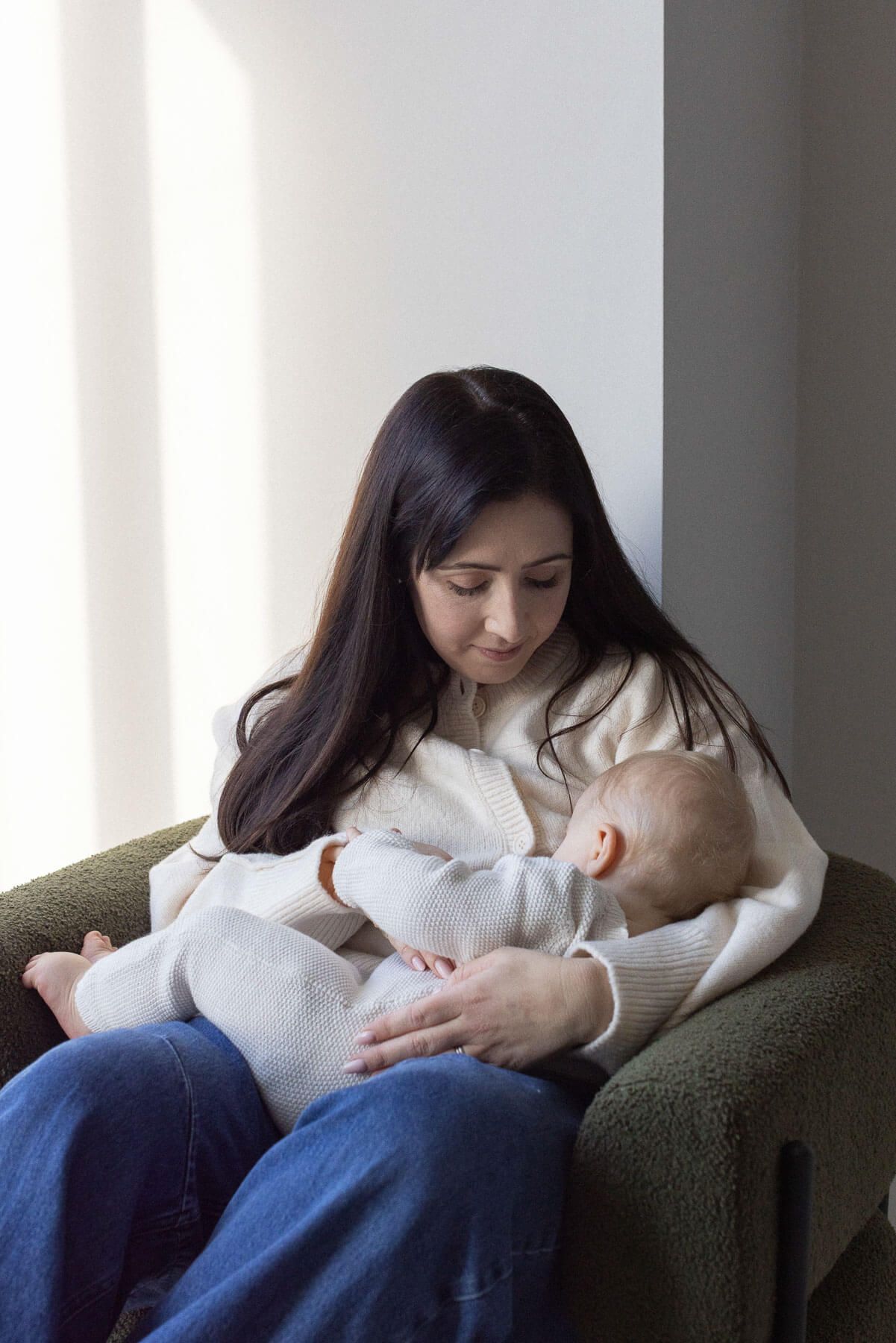 Mother sitting in a chair breastfeeding her baby in a bright, naturally lit room, photographed during an in home session in Brighton